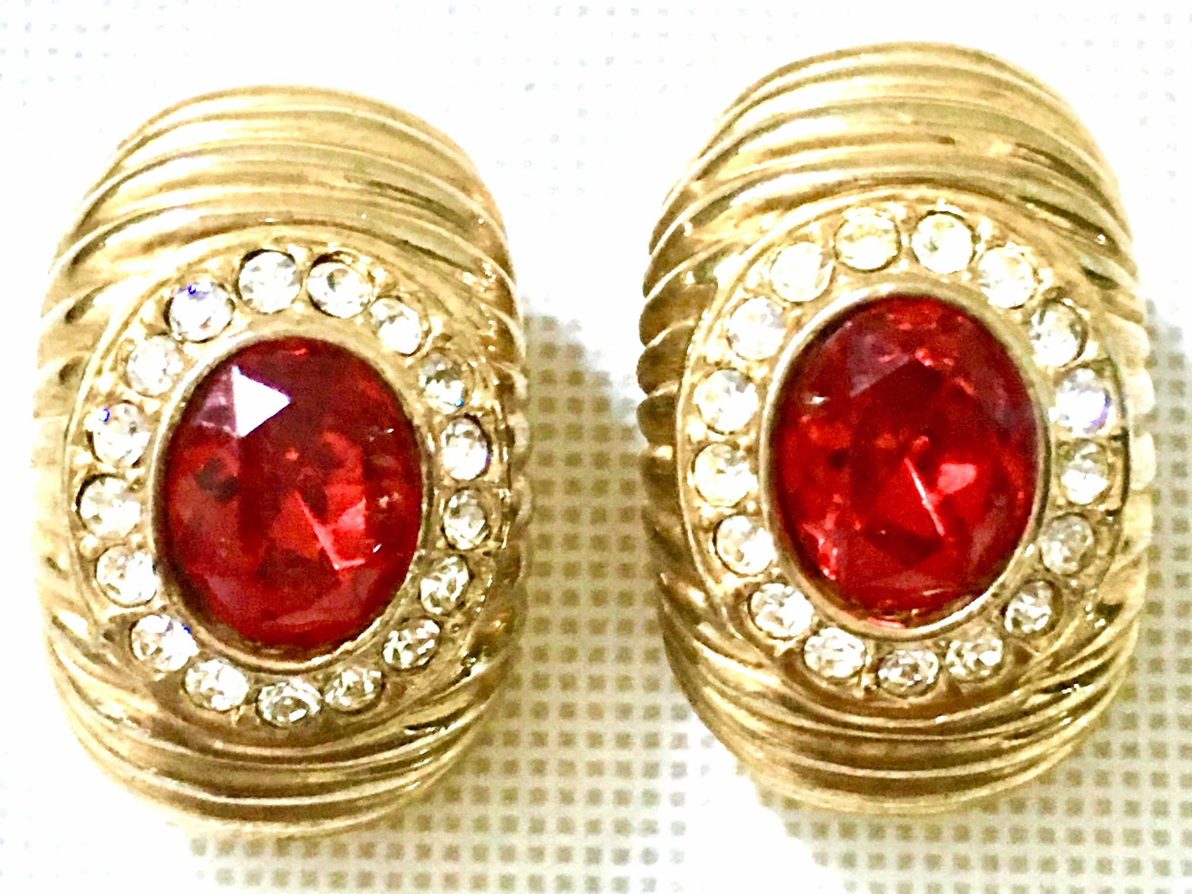 20th Century Gold & Swarovski Earrings By, Christian Dior. These classic and timeless earrings feature a curved and dimensional shape. Executed in gold plate metal with ridge detail and a large central ruby red cabochon stone surrounded by colorless