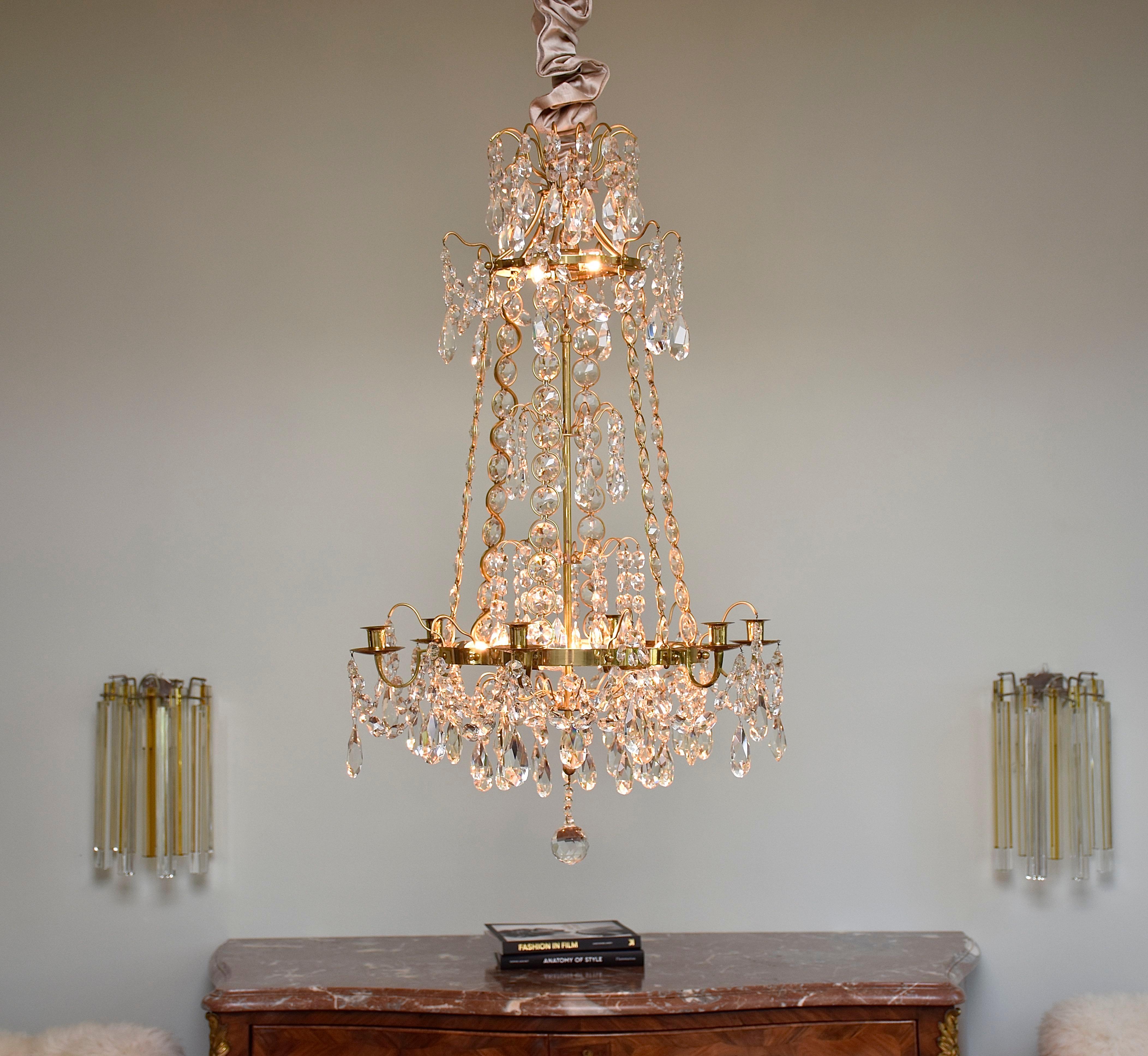 Un lustre élégant et fin de style gustavien avec 8 petites lumières à l'intérieur du lustre.
Belle qualité en laiton doré.
Décoré de cristaux taillés, de gouttes, de boutons et de chaînes. Six chaînes reliant l'anneau de la couronne à l'anneau