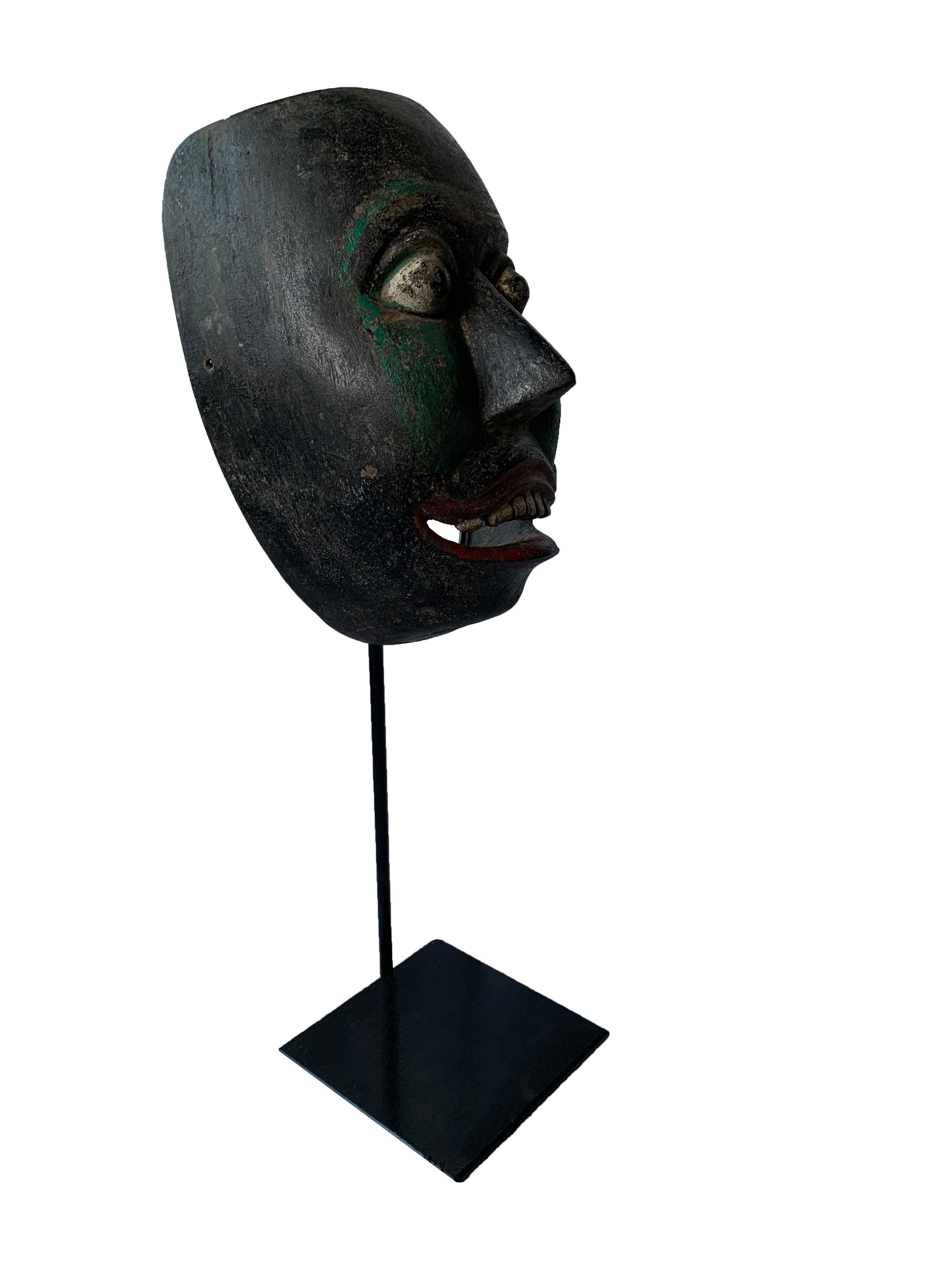 This early 20th century ‘Wayang topeng’ mask was used in Javanese masked dance performances. This mask features a black and green polychrome finish on its face with burgundy lips and upper teeth exposed. Much of the polychrome has faded over the