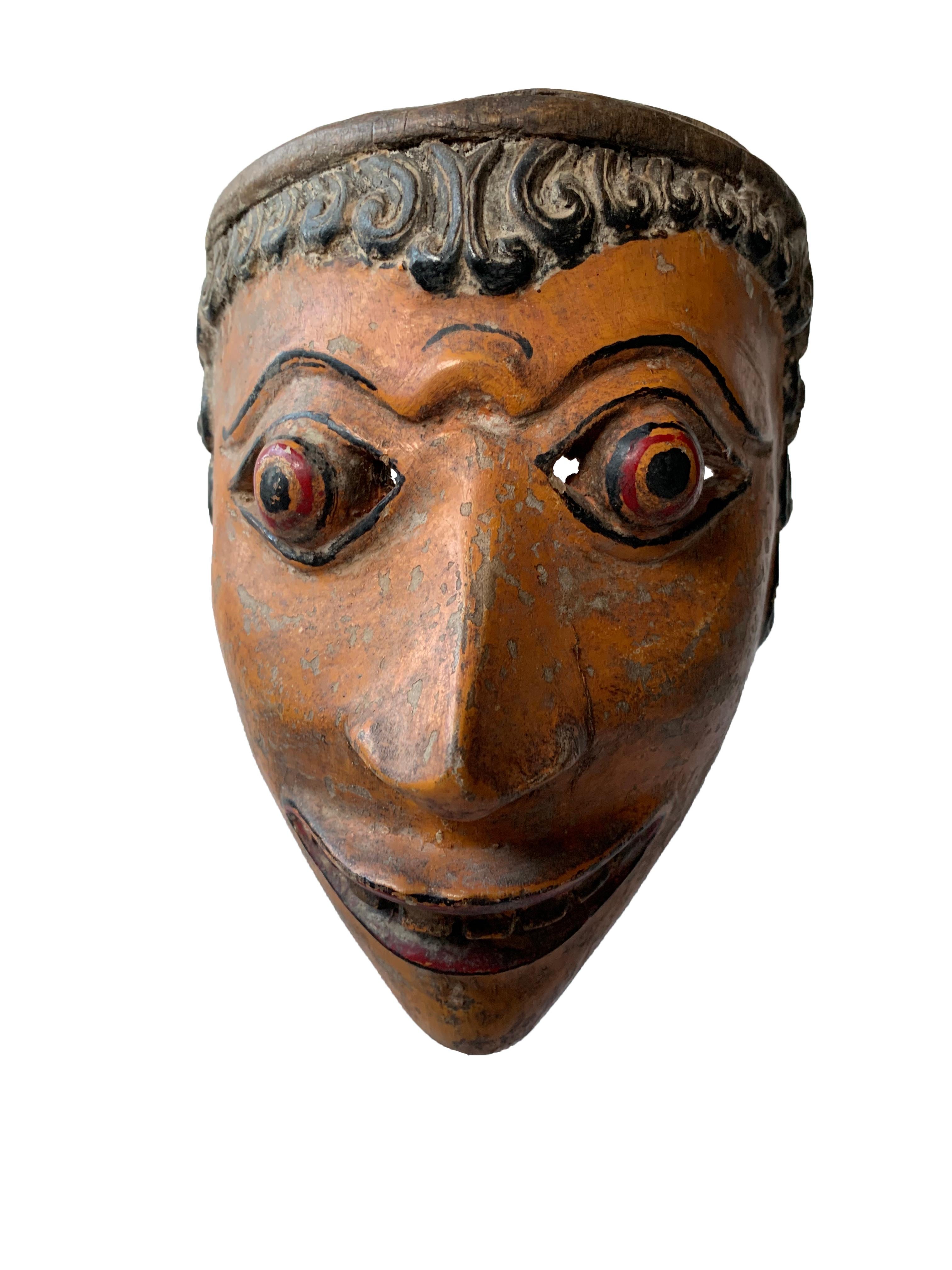 This early 20th century ‘Wayang topeng’ mask was used in Javanese masked dance performances. This mask features an orange polychrome finish with red coloured eyes that form a dramatic appearance. 

Measures: Height with stand 48cm; Height 19cm x