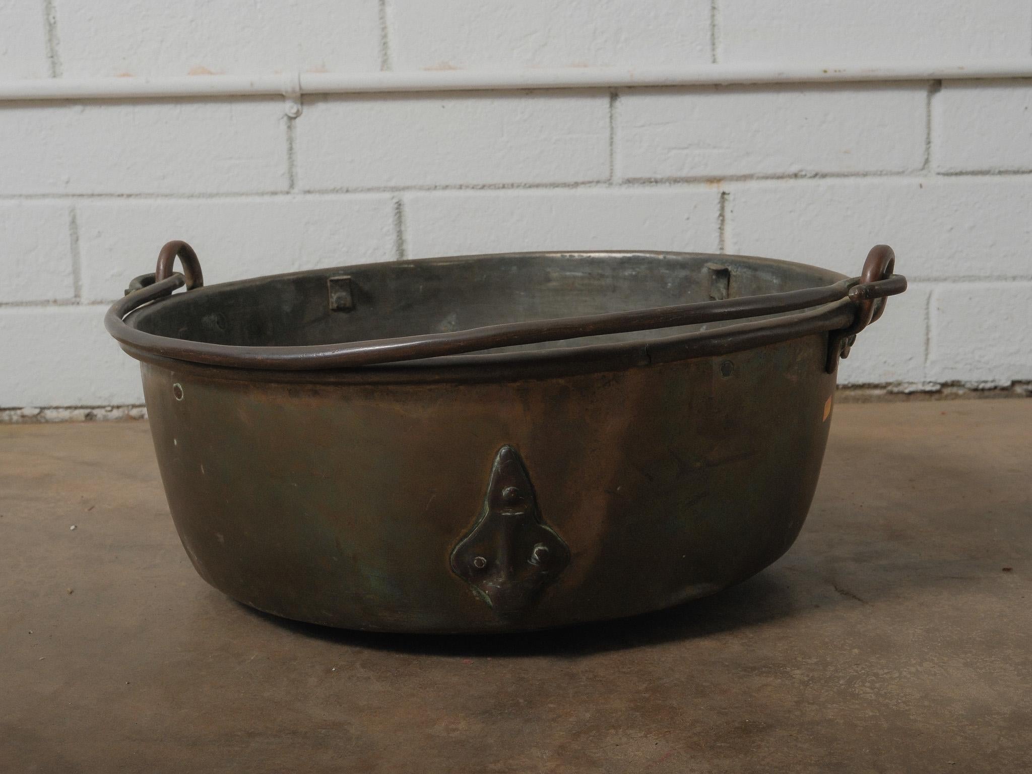 A substantial hand-forged copper vessel, defined by its broad proportions, sculptural hardware, and a deep, time-softened patina. The piece features a single swinging iron handle that arcs across the top, secured by forged brackets on each side. A