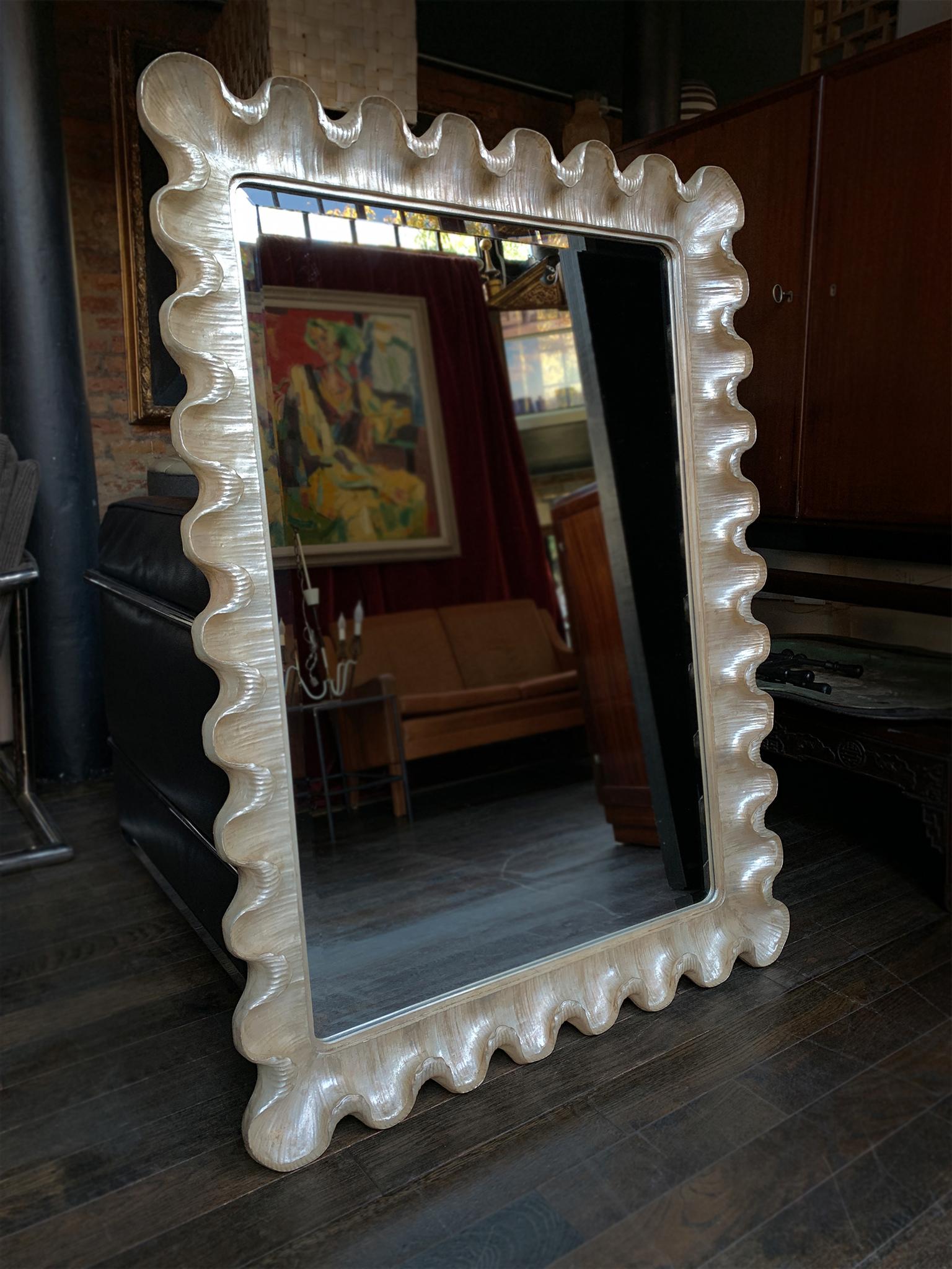 A vintage wall mirror in a lush Hollywood Regency style. We love the frame's beautifully carved and scalloped form, which remind us of delicate seashells. It is painted in a champagne color with a pearlescent shine. The glass is beveled and secured