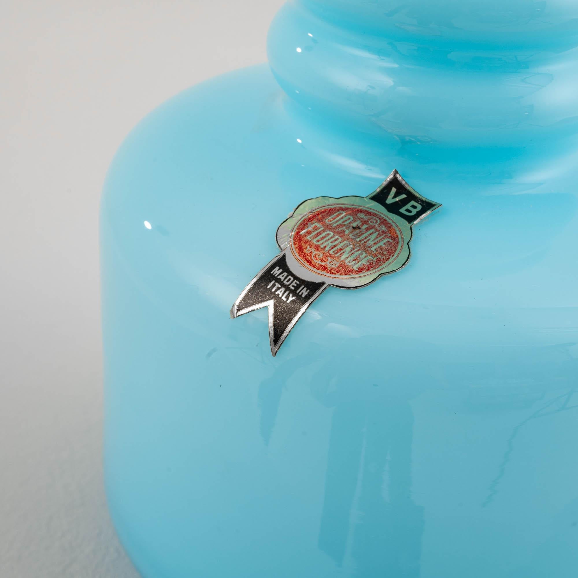 Our 20th Century Italian Opaline Blue Glass Jug is a striking embodiment of elegance and functionality. With a wide base that gently narrows, accentuated by two delicate rings, the jug exudes a balanced and harmonious design. The ice-blue hue adds a