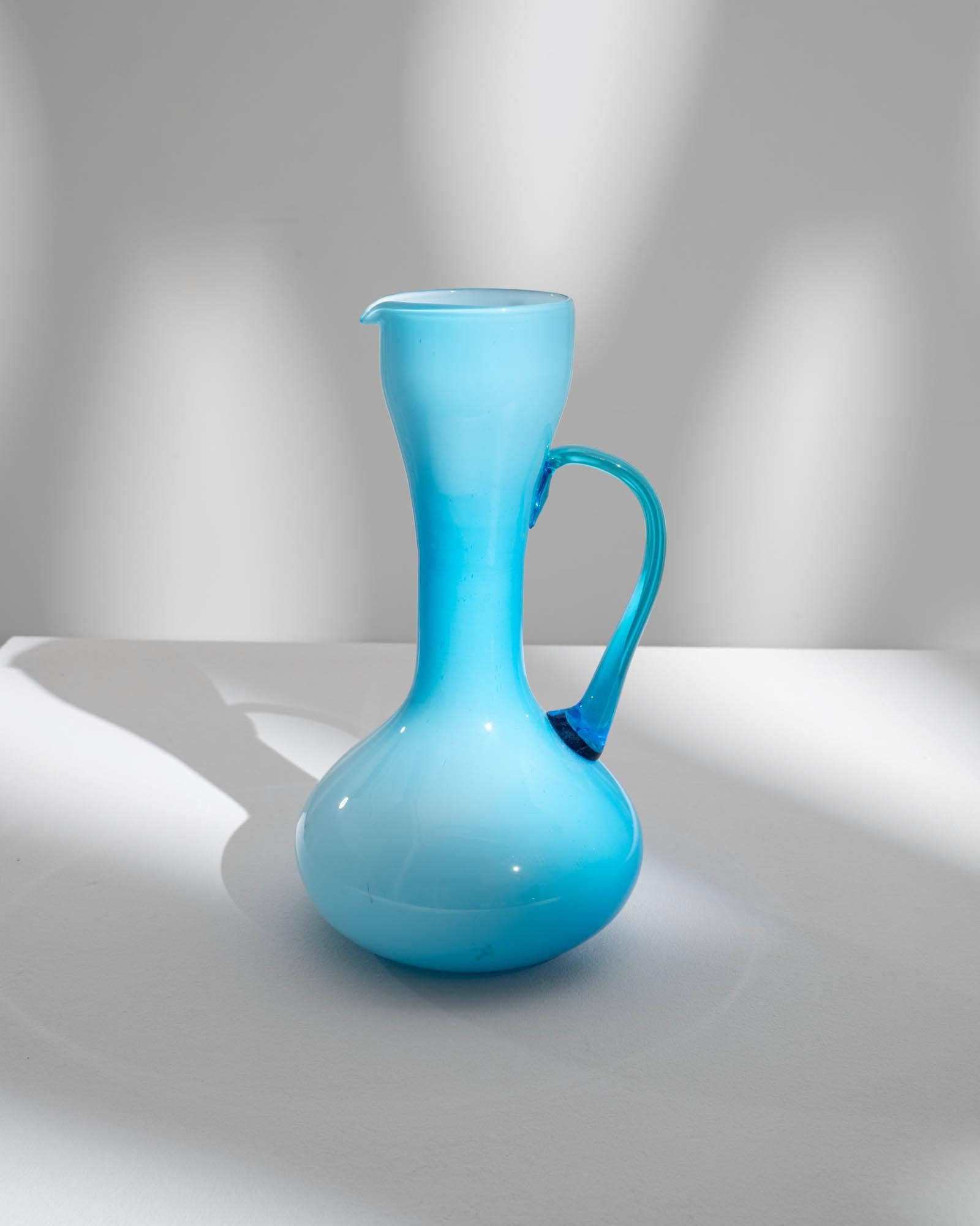This exquisite 20th Century Italian Ice Blue Glass Jug is a vision of sophistication. The striking ice blue color, reminiscent of frozen tranquility, graces the jug's wide base before elegantly tapering towards the top. The smaller, wide opening is