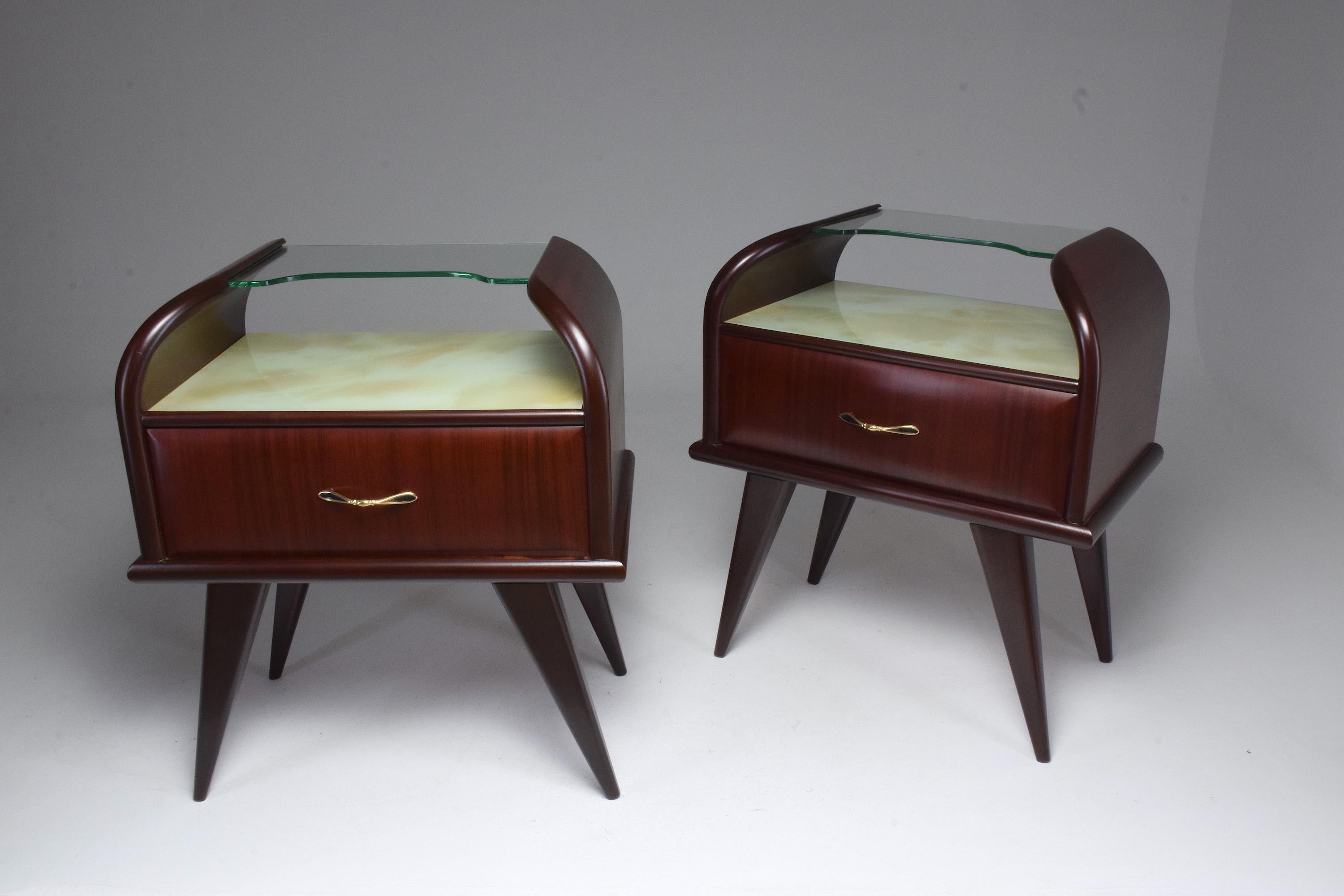 A pair of Italian midcentury nightstands or bedside table in mahogany designed with two glass shelf units, a central drawer with a sophisticated gold polished brass handle and splayed tapered legs typical of the Italian design period. The main shelf