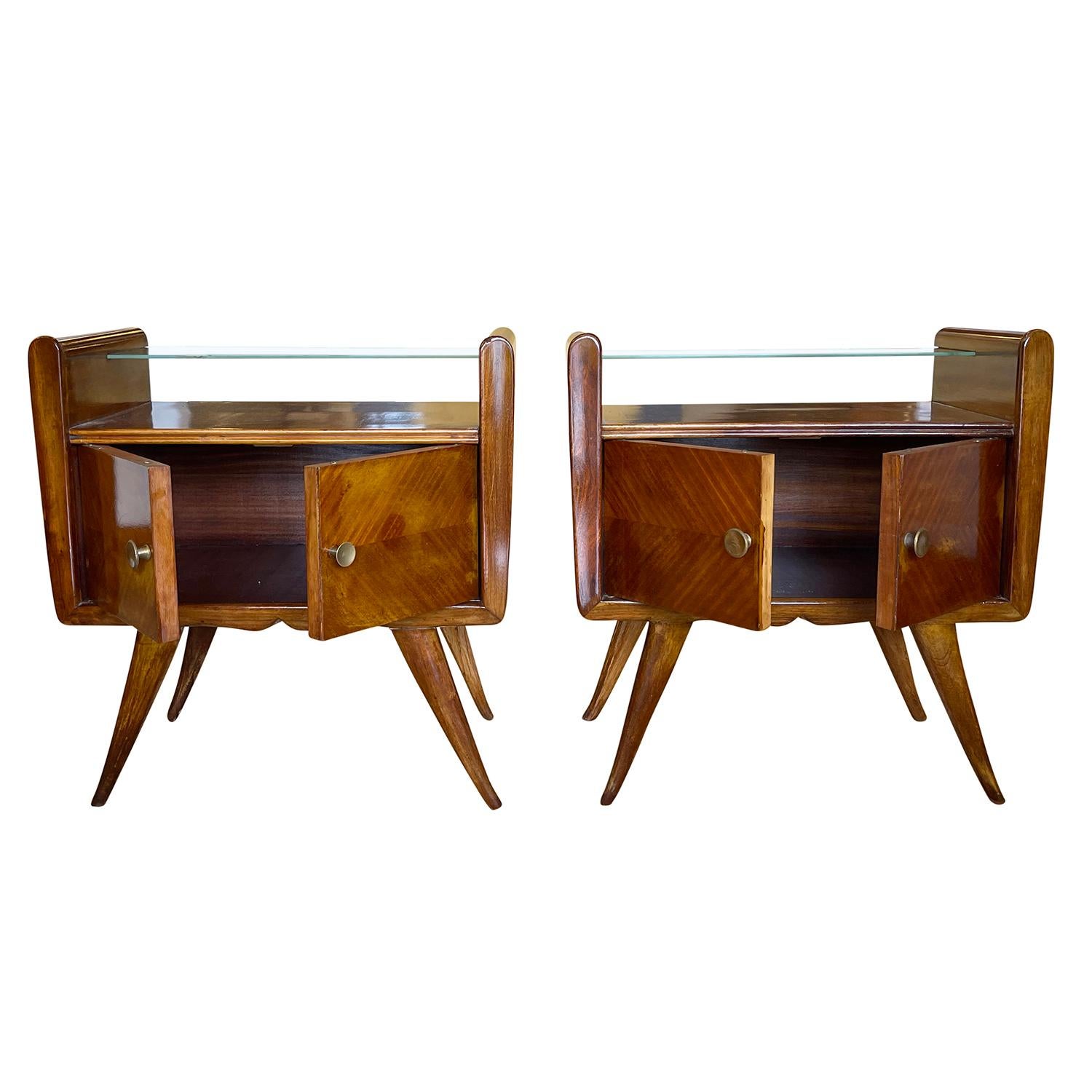 A vintage Mid-Century Modern Italian pair of nightstands made of hand carved walnut with slotted clear glass half shelf, composed with two cabinet doors and one varied, brass door pull, in matched veneers. The bedside tables are standing on four