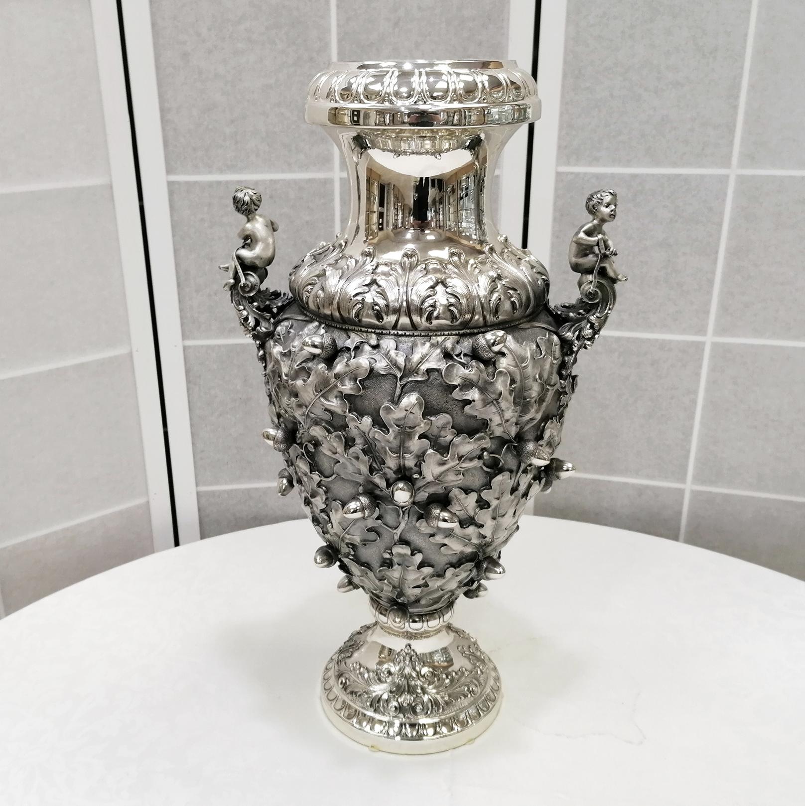 Wonderful big vase with shaped round body embossed, chiselled and engraved with oak leaves.
On the body are soldered melting made acorns
Above the handles are placed two children.
The round base is also embossed and chiselled

The maker was a