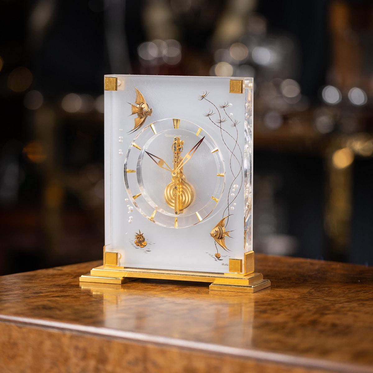 A Mid-Century mantel clock by Jaeger-LeCoultre graces its surroundings with an elegant Lucite case adorned with inset brass decorations depicting fish among reeds and rising air bubbles. It operates on an eight-day movement, featuring