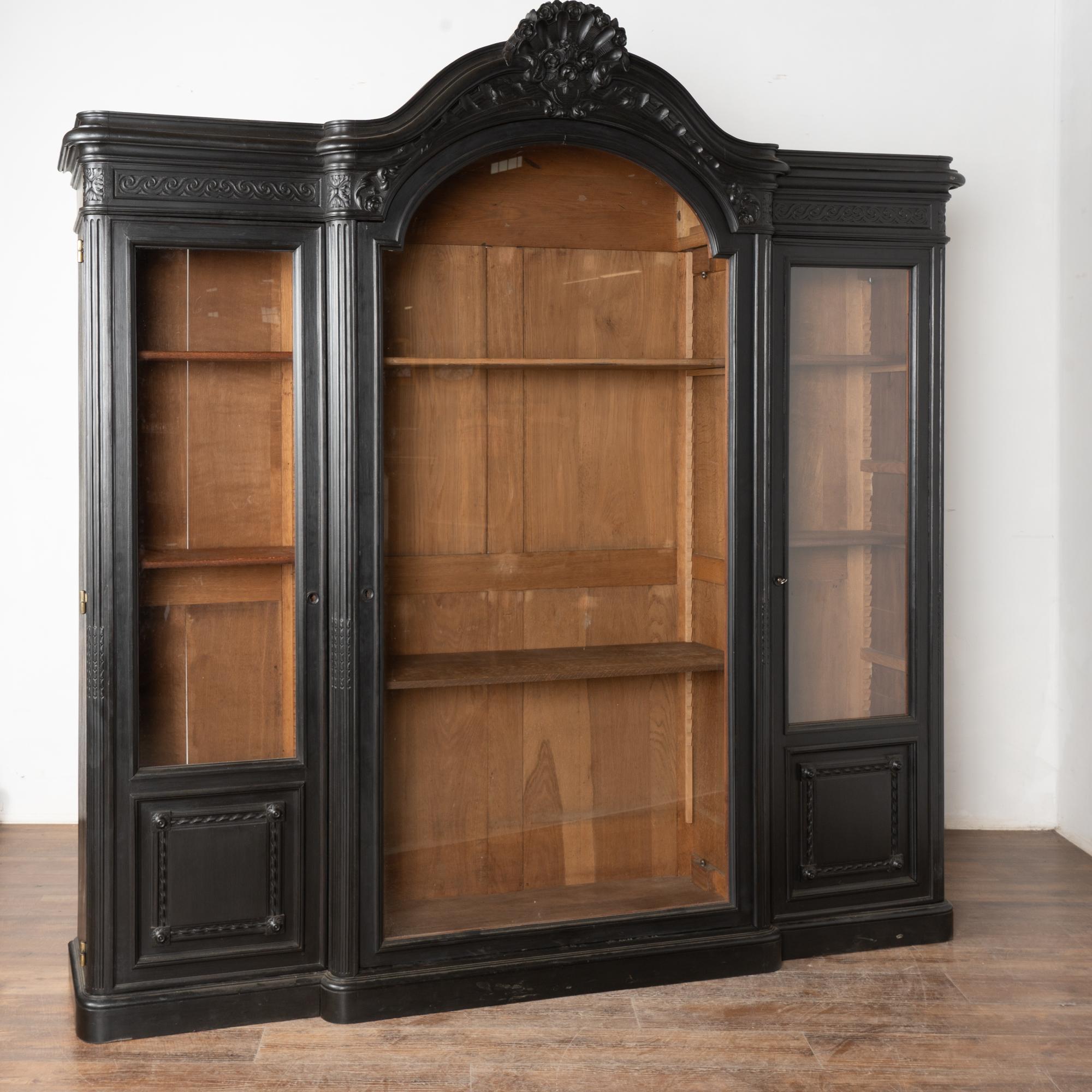 Questa grande libreria o vetrina in Oak ha tre lunghe ante in vetro e supera i 7,5' di altezza e larghezza.
I dettagli intagliati, tra cui rosette e altre decorazioni, impreziosiscono i pannelli inferiori, i lati e la corona centrale