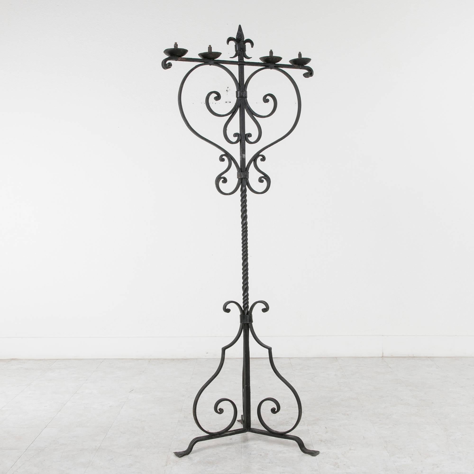 This early 20th century hand-forged iron torchere with fleur de lys accommodates four 3 inch diameter candles. The hand-forged iron tripod base with scrollwork supports a twisted iron stem that rises to a point created by the fleur de lys. This