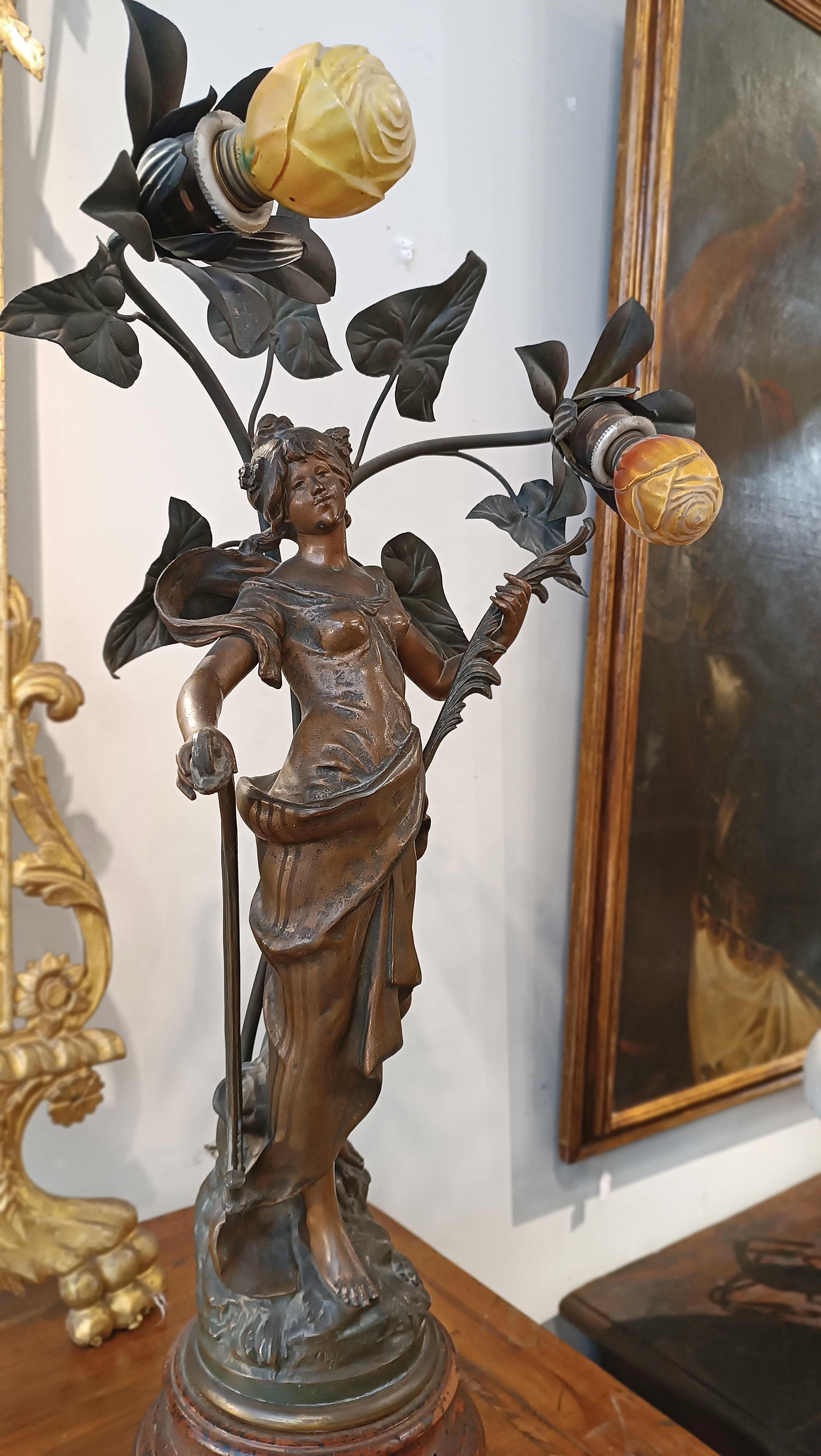 This Liberty lamp in bronze alloy represents a true work of decorative art. The female figure statuette is striking for its grace and delicacy. The woman holds a harp, symbol of harmony and purity, and a palm, which represents victory and nobility
