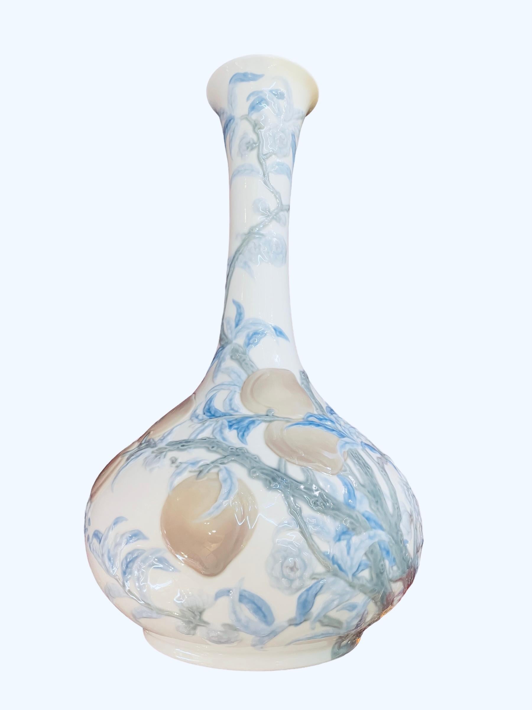 This a Lladro soft glow porcelain flower gourd vase. It is hand painted white in the background with a repousse of long light green branches with light blue leaves and flowers plus rose-orange color peaches hanging from them. Under the base is