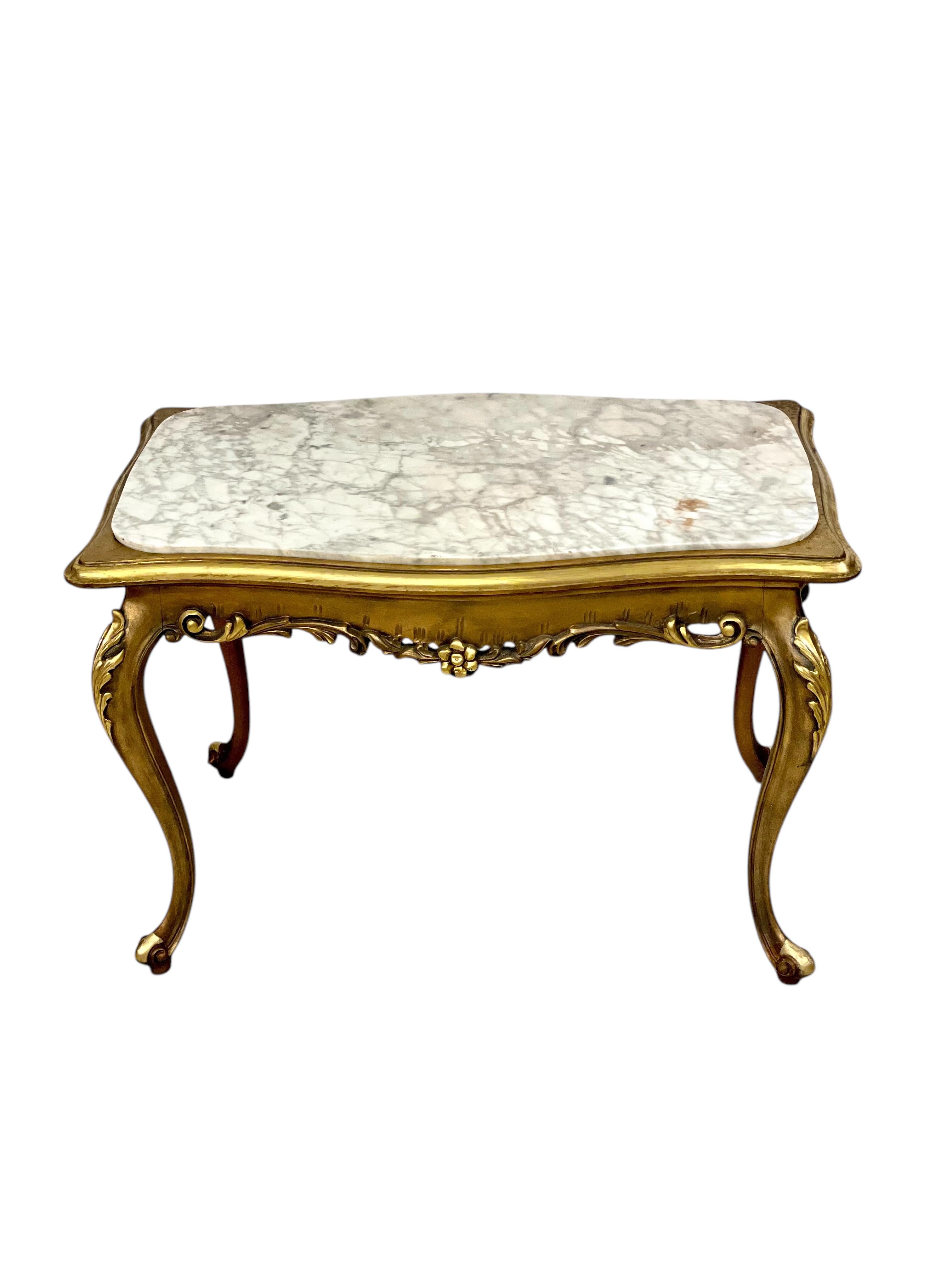 Cette élégante table basse française du début du XXe siècle incarne le style rococo raffiné inspiré du design Louis XV. La table présente un cadre en bois doré sculpté et orné de pieds cabriole sinueux, chacun agrémenté de délicats motifs floraux et