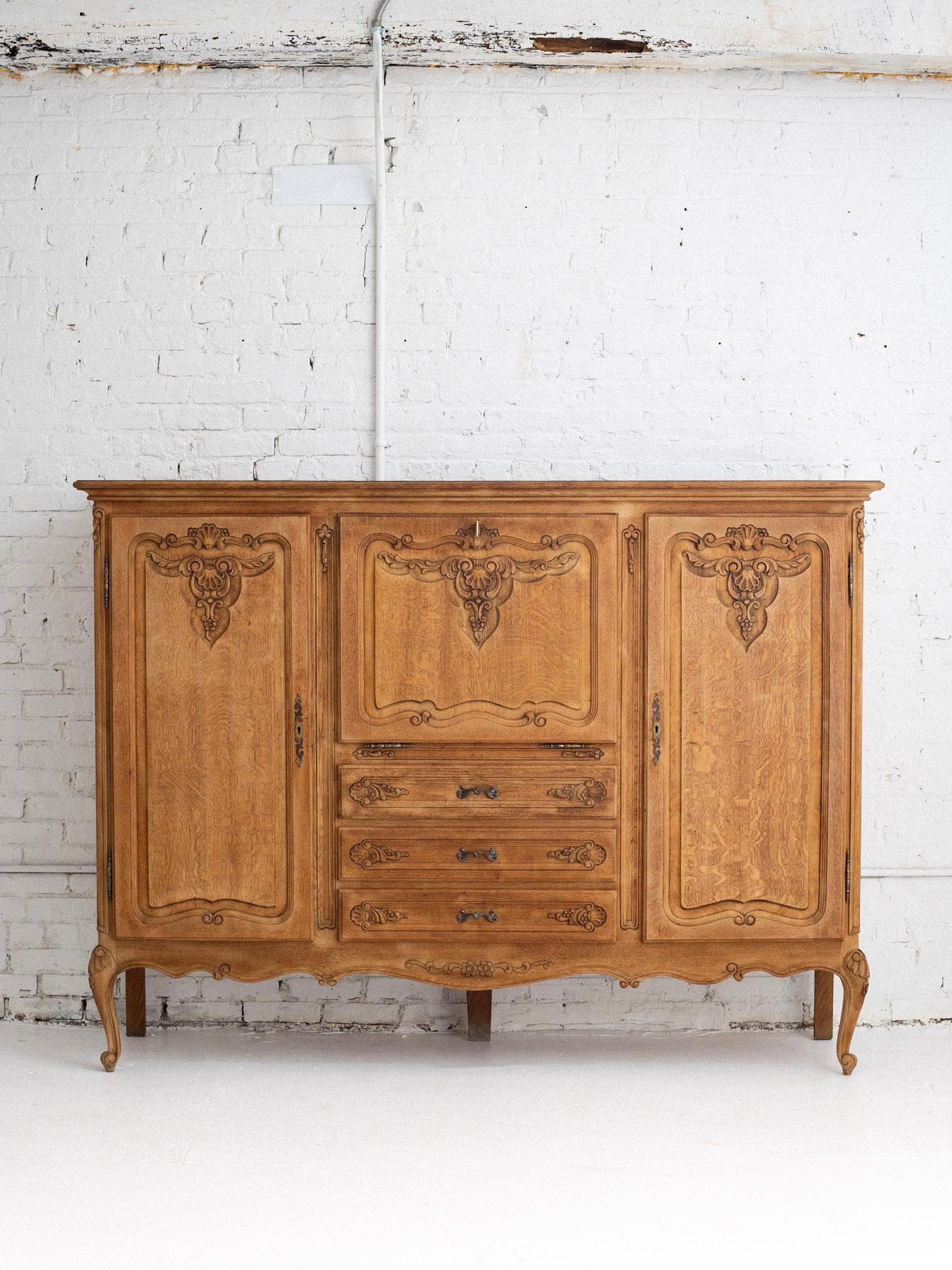 Mueble bar francés de mediados del siglo XX de estilo Luis XV. Un acabado rústico de roble blanqueado. Dos armarios exteriores ocultan la estantería interior. Tres cajones centrales se sitúan debajo de un mueble bar abatible con estante de cristal
