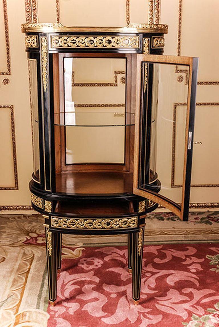 French salon vitrine in Louis 16th classicist style.
Piano black polished veneer on solid pinewood. Oval one- drawered combered
and four-sided glassed body on high, Brasse-painted four edged legs,
ending in Sabots. Border and cornicing with
