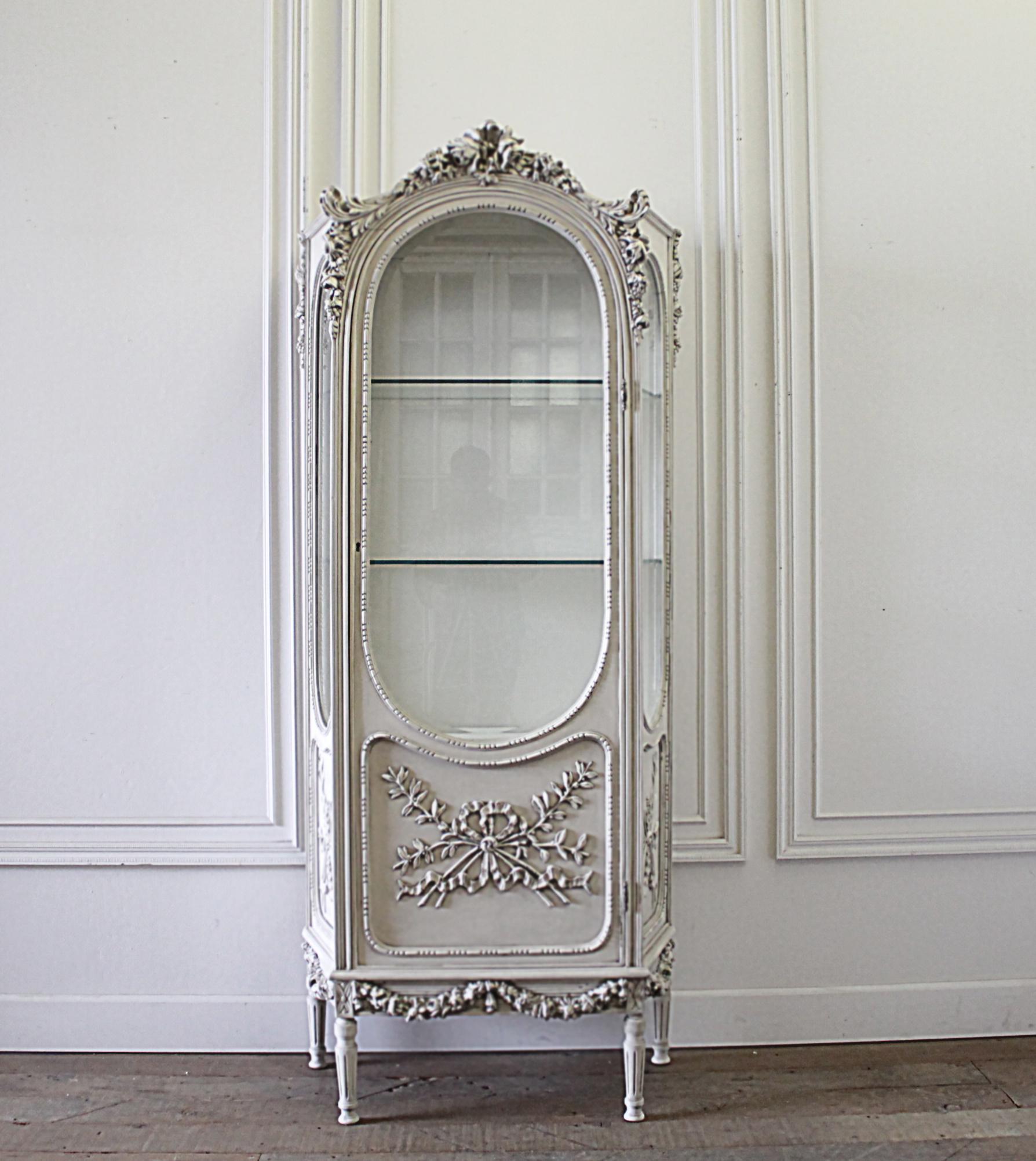 20th century Louis XVI style carved display armoire curio with rose carvings
Painted in our beautiful antique white with subtle distressed edges and finished with an antique patina. Beautiful carved rose details, and fluted legs. There are 2 glass