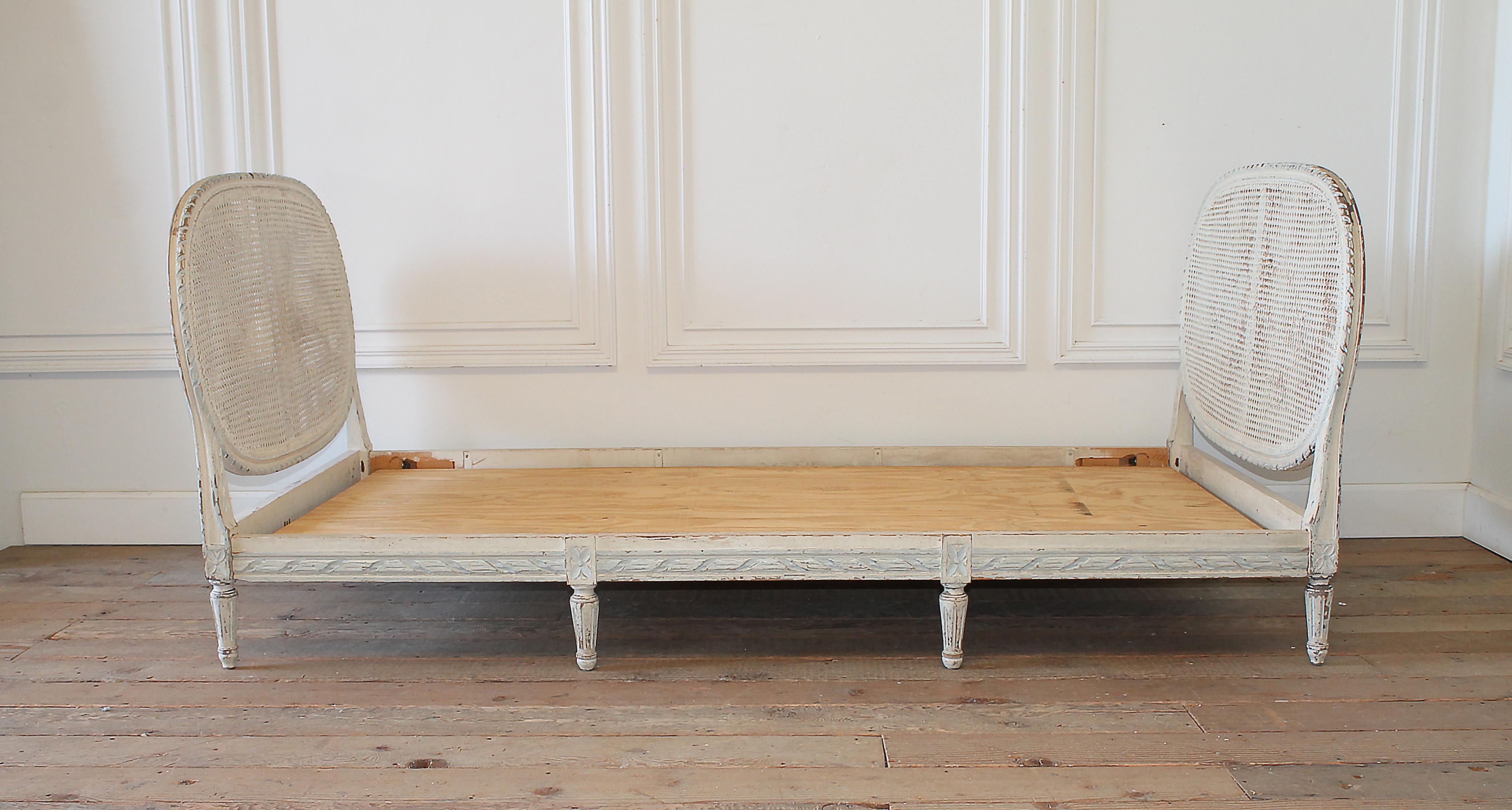 20th century Louis XVI style original painted French cane daybed
Paint is in off-white with antique distressed edges, accent paint colors on carving is a pale grey blue, cane is in good condition. No breaks or damage. The cane has a chippy paint