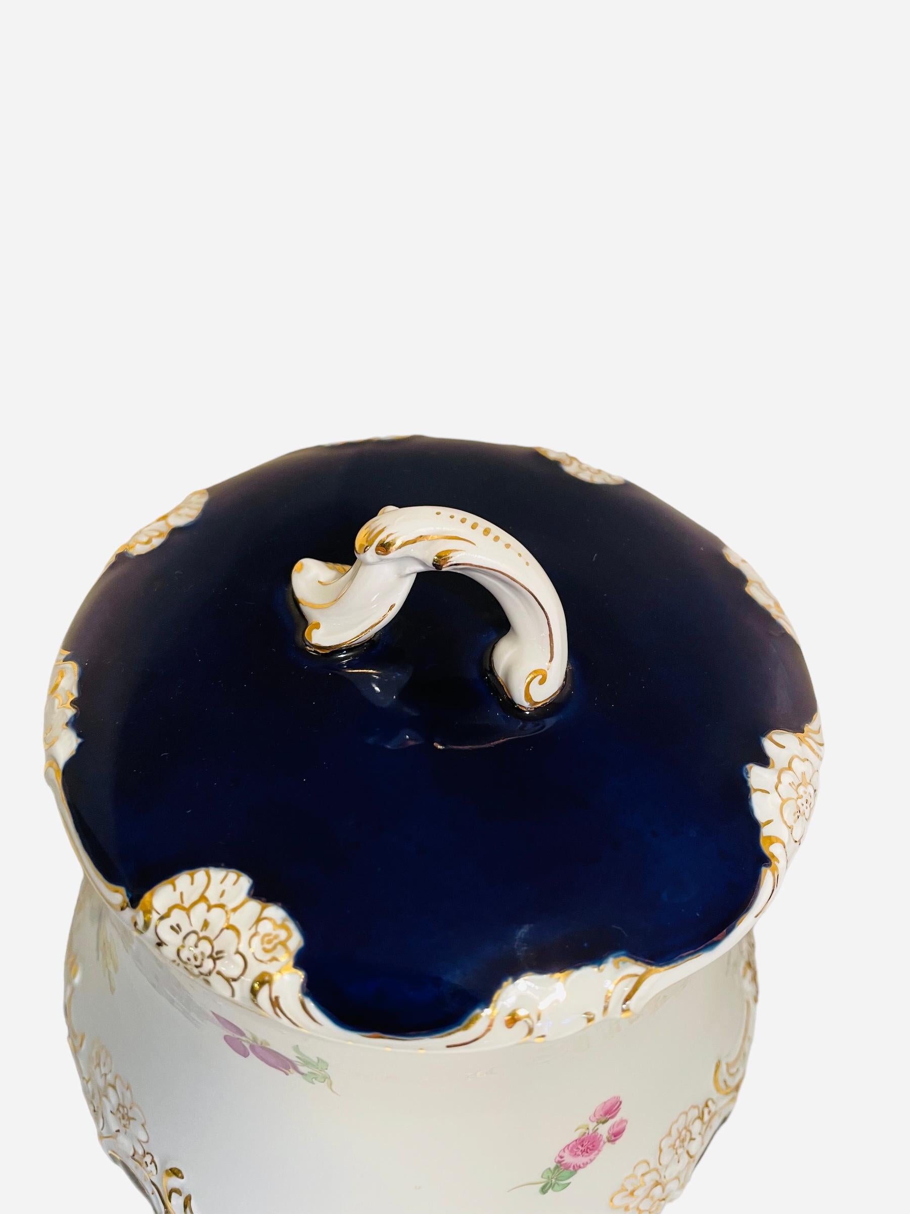 This is a 20th century Meissen porcelain cookie/ginger lidded jar. It depicts a barrel shaped jar hand painted white in the background and cobalt blue in the lid and bottom area. Small bouquets of different flowers adorn the body of the jar. A