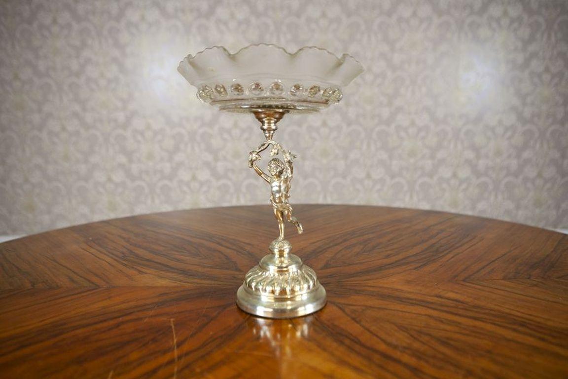 20th-Century Metal Fruit Bowl With Cherub

Metal fruit bowl with a foot in the form of a cherub holding a glass bowl of the vessel.
Minor imperfection on the glass (see picture)