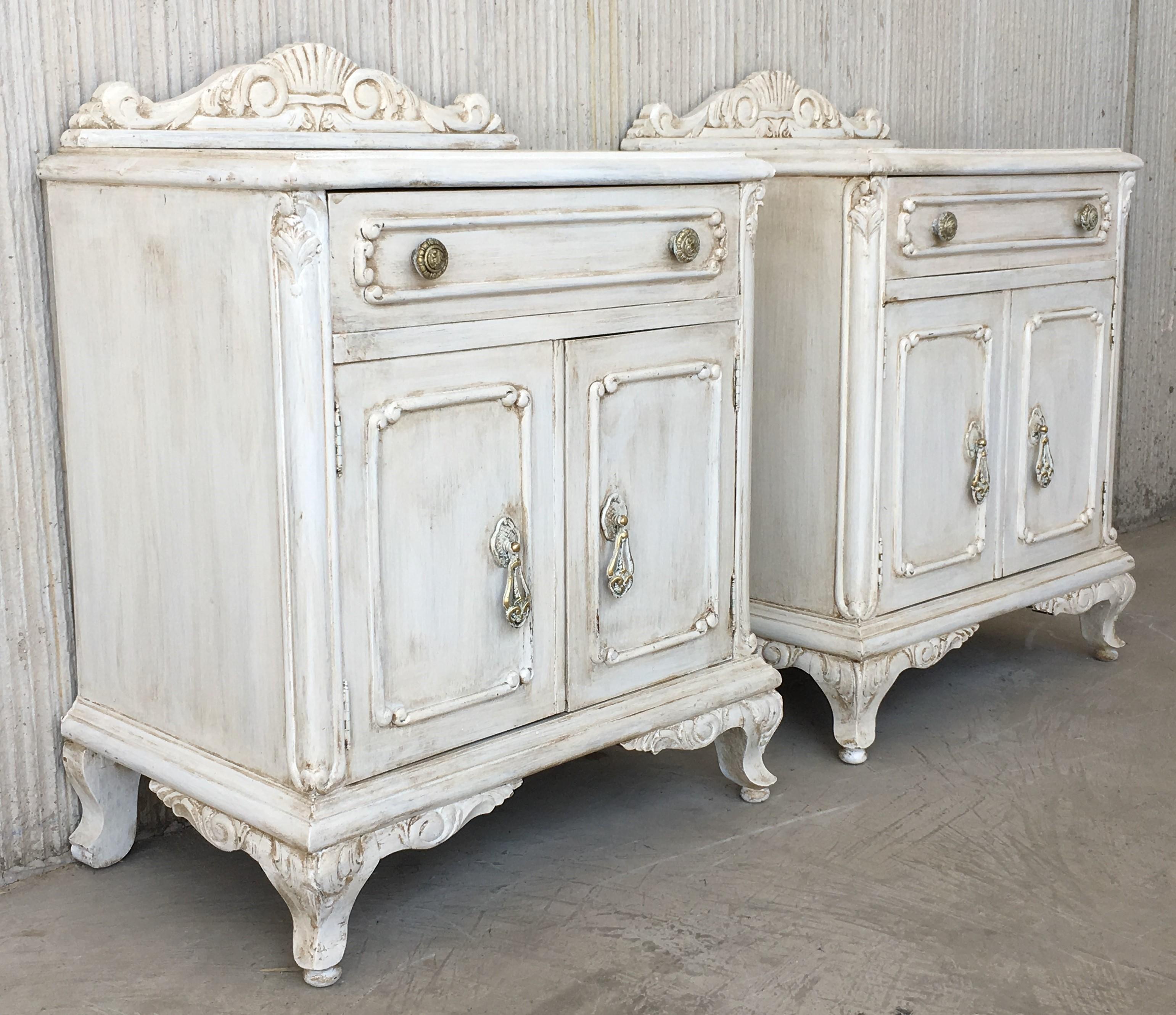 20th century vintage neoclassical nightstands with crests in white patina with drawer and two doors.

Pair of vintage neoclassical style nightstands painted in a subtle oyster white finish. Off-white, with distressed edges, and antique glazed