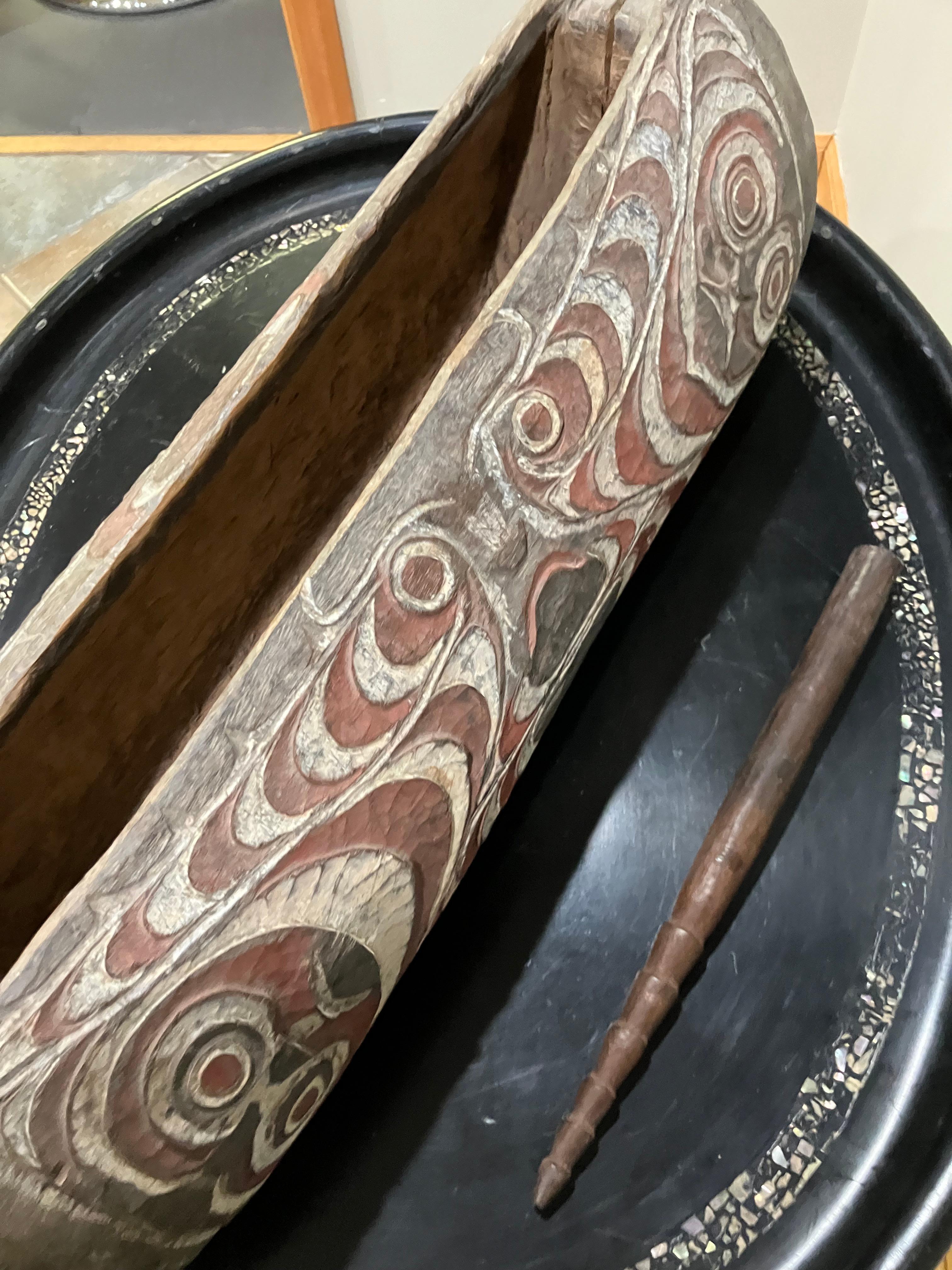 Hand-carved wooden drum from New Guinea, featuring intricate painted patterns in white, red, and black across the cylindrical body. The surface displays symmetrical curvilinear motifs, with stylized face-like elements at each end. The drum includes