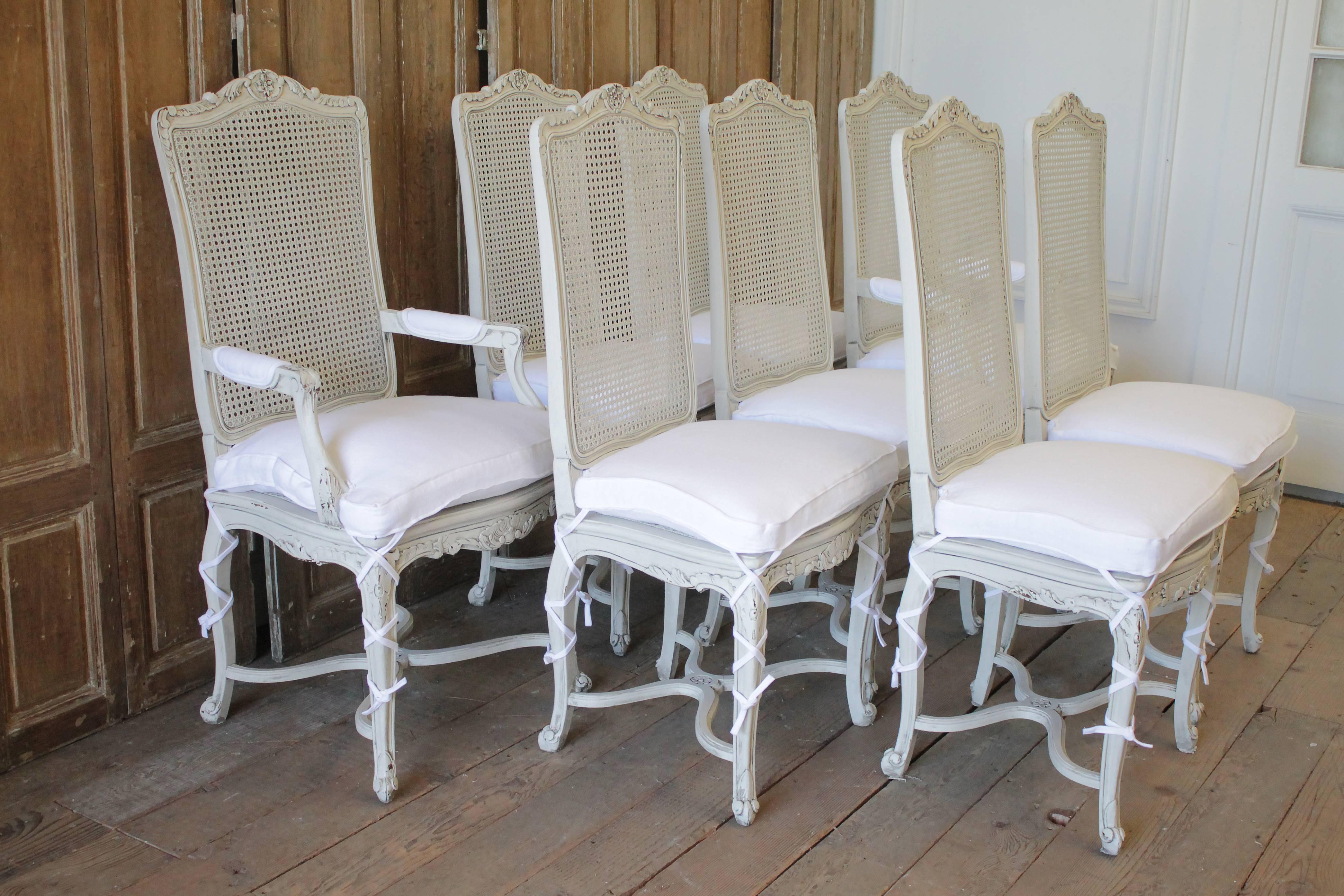 20th century painted Louis XV style cane back dining chairs with linen cushions
Freshly painted in a soft white with subtle distressed edges, and finished with a hand applied antique glaze to give the look of a time worn patina. Cane is good