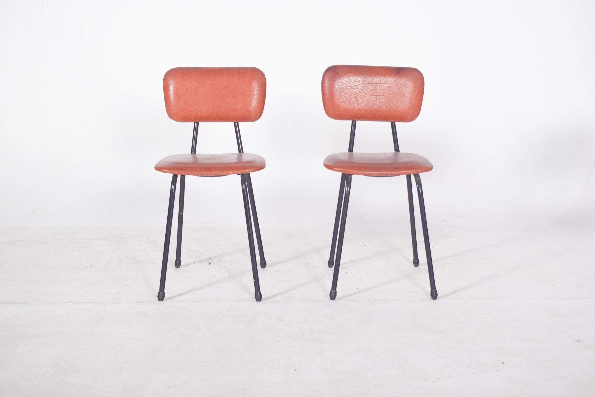 Pair of chairs in red synthetic leather. With a black tubular metal frame and upholstered seat and back, typical of 20th century school or office environments. The simple, functional lines are reminiscent of industrial design, prioritising