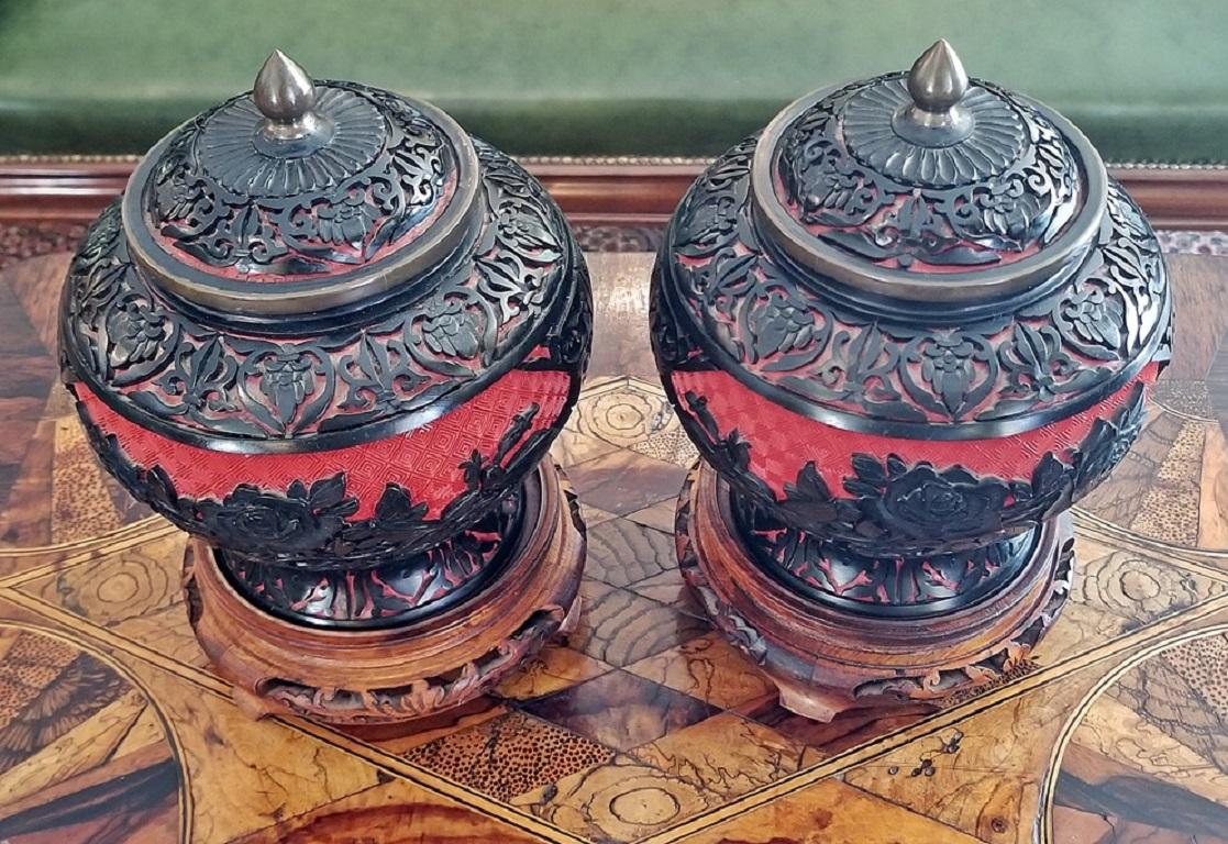 Gorgeous pair of early 20th century Chinese cinnabar lacquer and enameled lidded urns or vases on wooden stands.

Each urn consists of an enameled base (Cloisonné), blue enamel on the interior and red checkered geometric pattern enamel on the