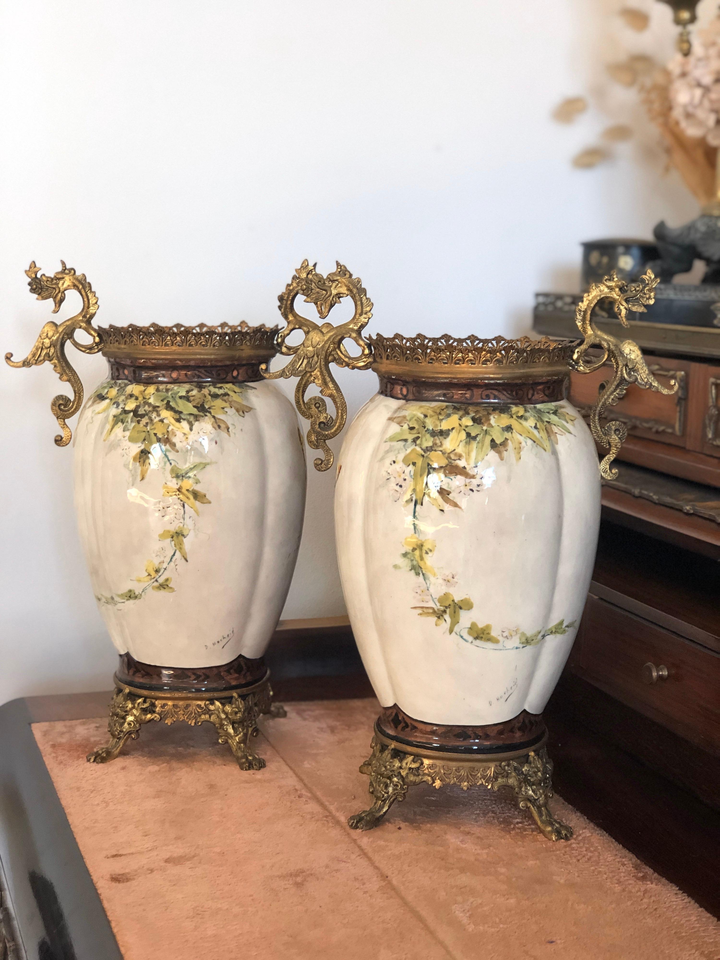 Pair of hand painted asymmetrical ceramic vases decorated with flowers and butterflies in light colors on beige base, and they are painted in cobalt blue inside. Both are raised over brass legs and have brass crowns on the top and brass hendles