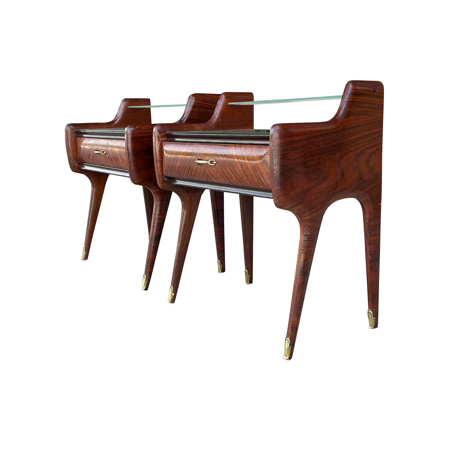 A dark-brown, vintage Mid-Century modern Italian pair of nightstands or side bed tables made of hand carved Rosewood with one-drawer and lacking glass shelves, enhanced by very detailed brass handles, supported on tapered legs. The ebonized bed side