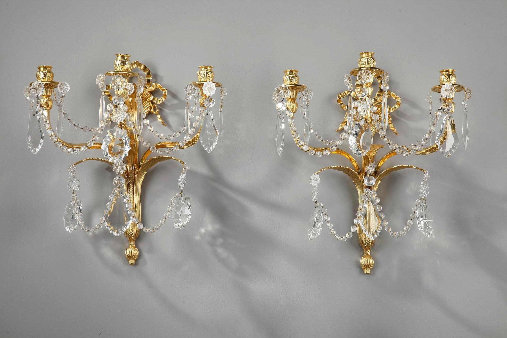 Pair of neoclassical wall lights sconces crafted of gilt bronze, with three-light. Each sconce is richly decorated with crystal drops, rosettes and drop-hung drippans. The ormolu fixture is chiseled with Classic Louis XVI decoration, including a