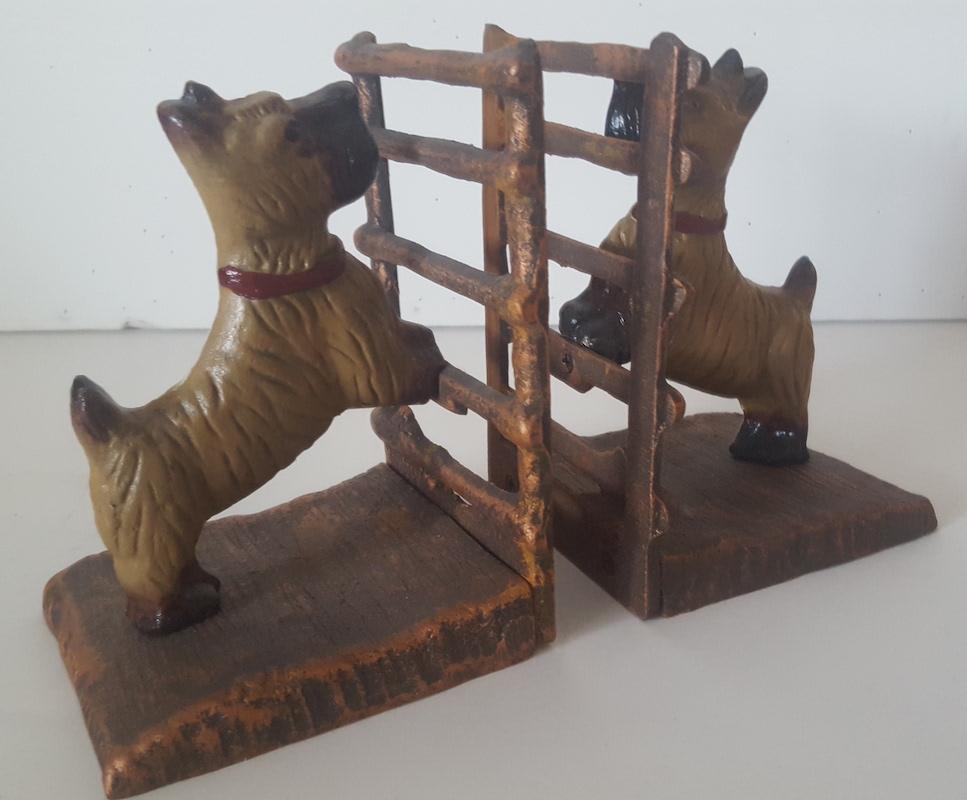 A matched pair of painted, cast dog bookends, circa 1930-1940, colored tan and black with red collars. Their front paws are up on the rung of a fence or gate which, along with the ground or base, is painted a metallic, patinated, copper or bronze