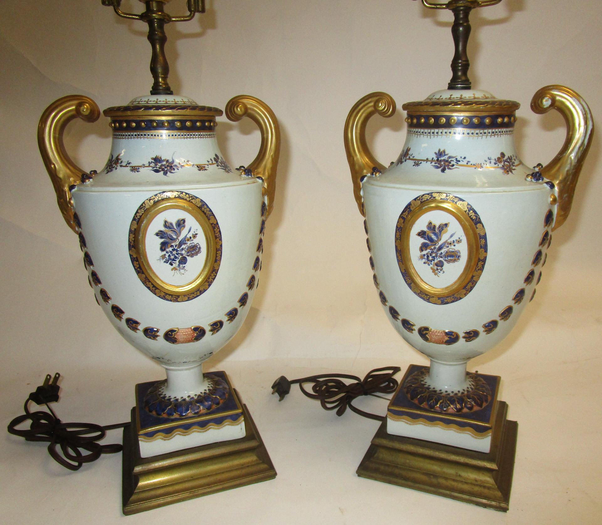 This handsome pair of Mottahedeh cobalt and white porcelain urn lamps feature central floral medallions surrounded by raised bellflower swags, hand-painted and parcel gilt, and are mounted on 5.5 inch square brass bases. Porcelain artichoke finials.
