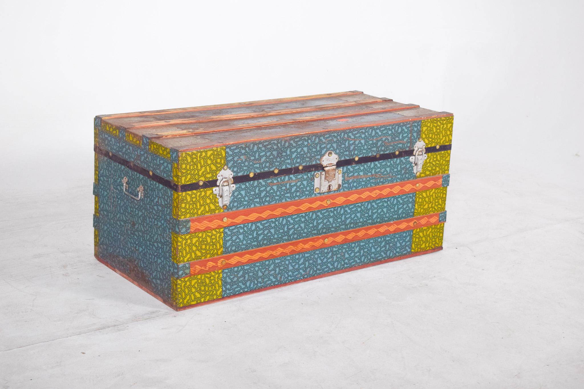 Baule in legno nei toni del verde, del blu e dell'arancione. Con una struttura robusta e decorata con vivaci motivi grafici, la parte anteriore della cassapanca è visivamente divisa da strisce verticali gialle alle estremità e da una sezione