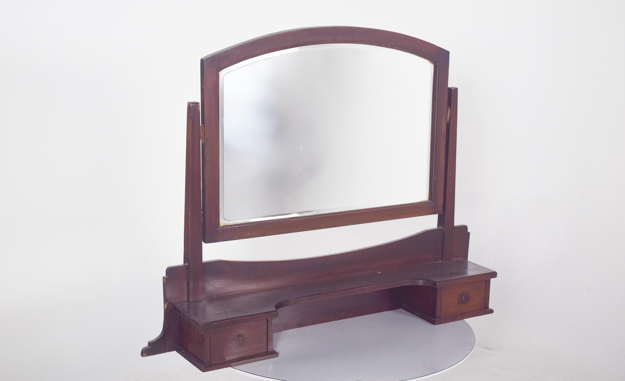 Specchio da toilette con due cassetti. In legno scuro, con una struttura robusta e un design sobrio. La cornice dello specchio ha una forma delicatamente arcuata nella parte superiore e lo specchio è fissato da due supporti laterali verticali, che