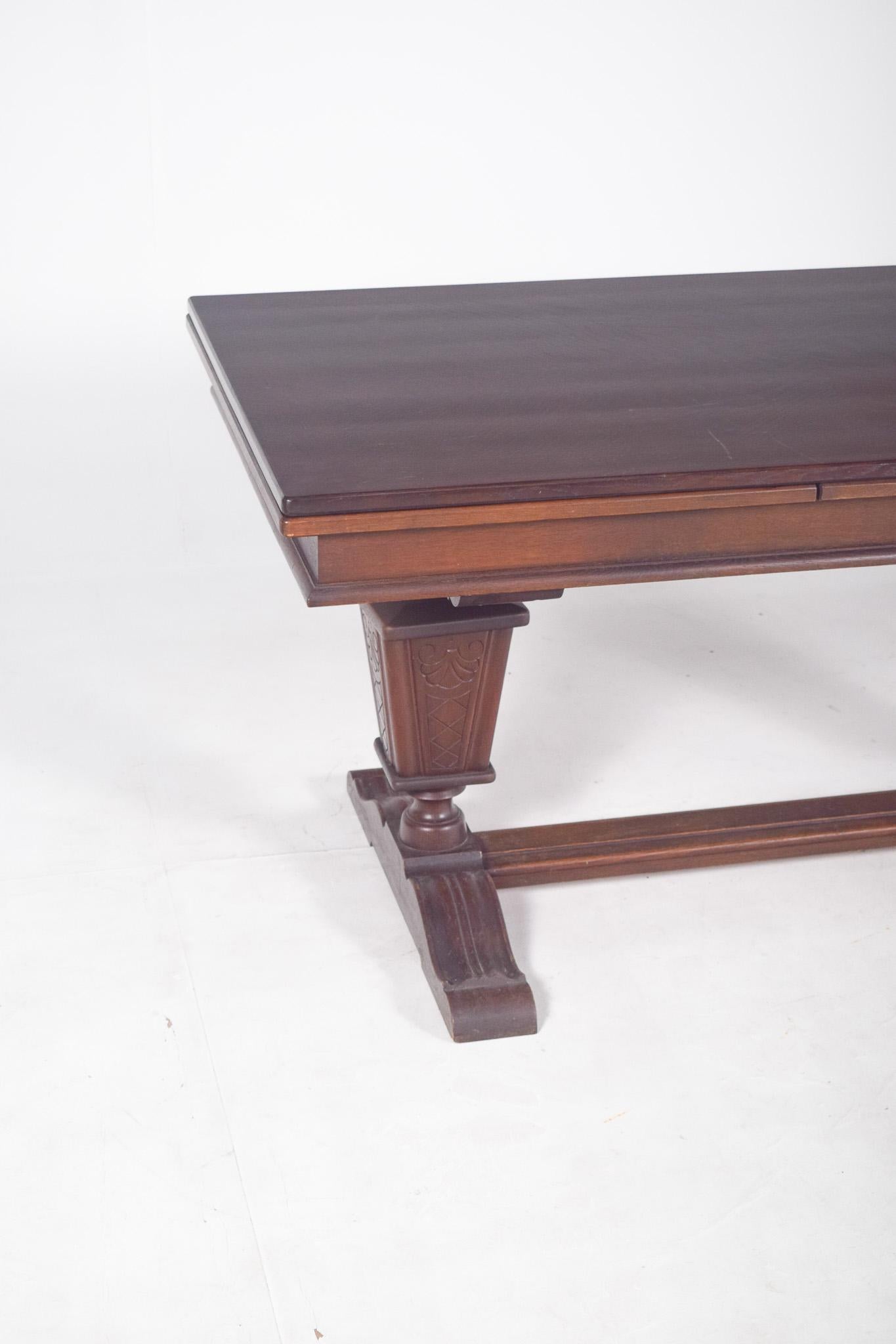 Tavolo da pranzo rettangolare allungabile. Il piano presenta bordi dritti con un profilo a gradini, mostrando un disegno geometrico caratteristico dello stile. Il supporto è composto da due sostegni centrali a forma di colonne, con intagli