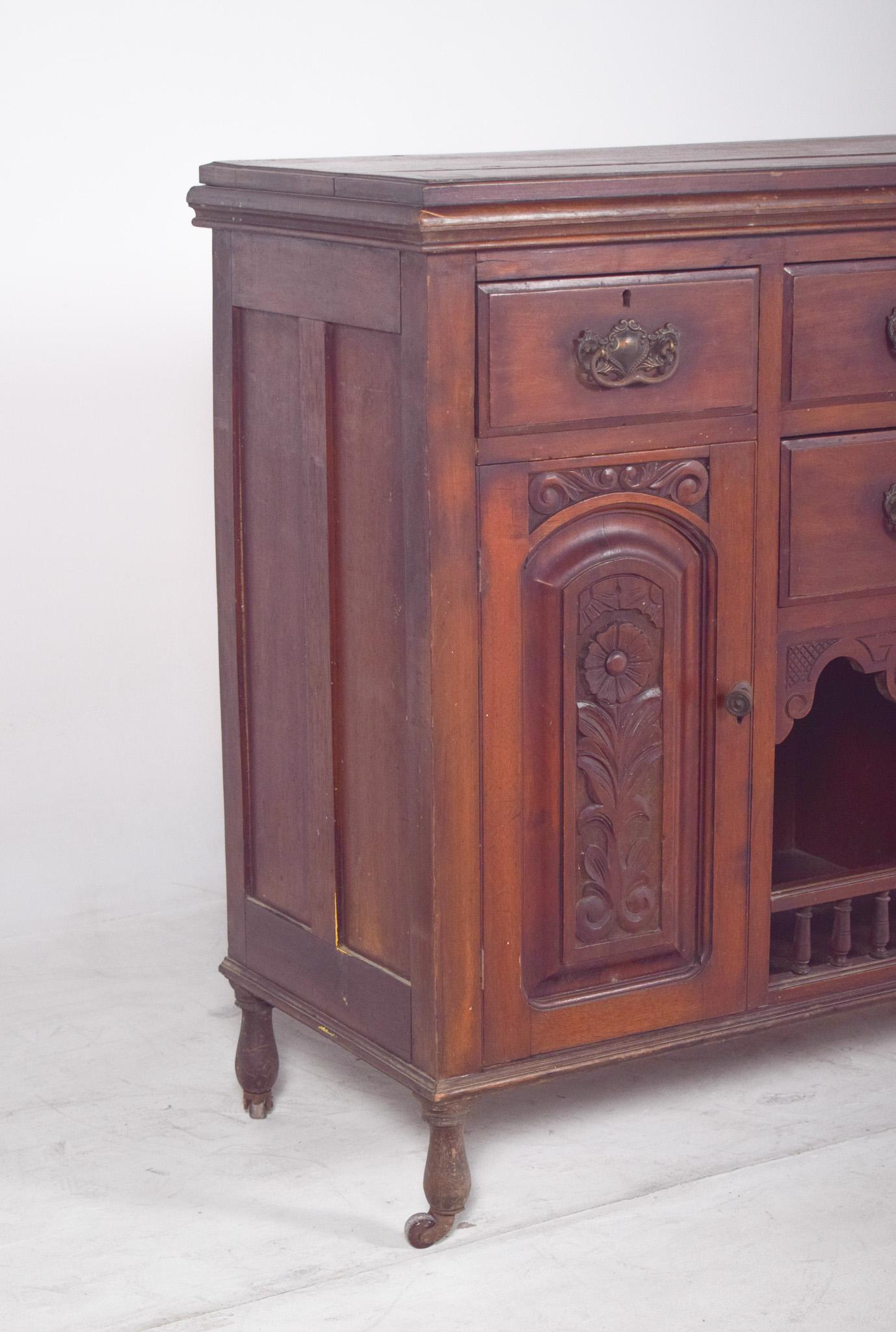 Aparador alto con dos puertas y cuatro cajones. Este aparador alto presenta un diseño robusto y funcional, típico de las piezas utilitarias de mediados del siglo XIX. De madera maciza, combina elementos decorativos tallados con una práctica