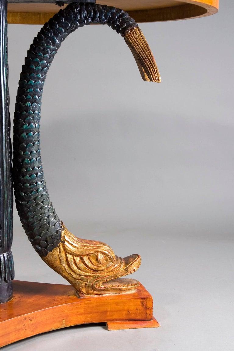 A classicist table with three monumental, full-plastic, hand carved dolphins made of solid wood. Scales colored, heads and fins are antique gold. Three-sided base plate on glass panes. Centrally ascending column, carved with leaves. Cover plate and