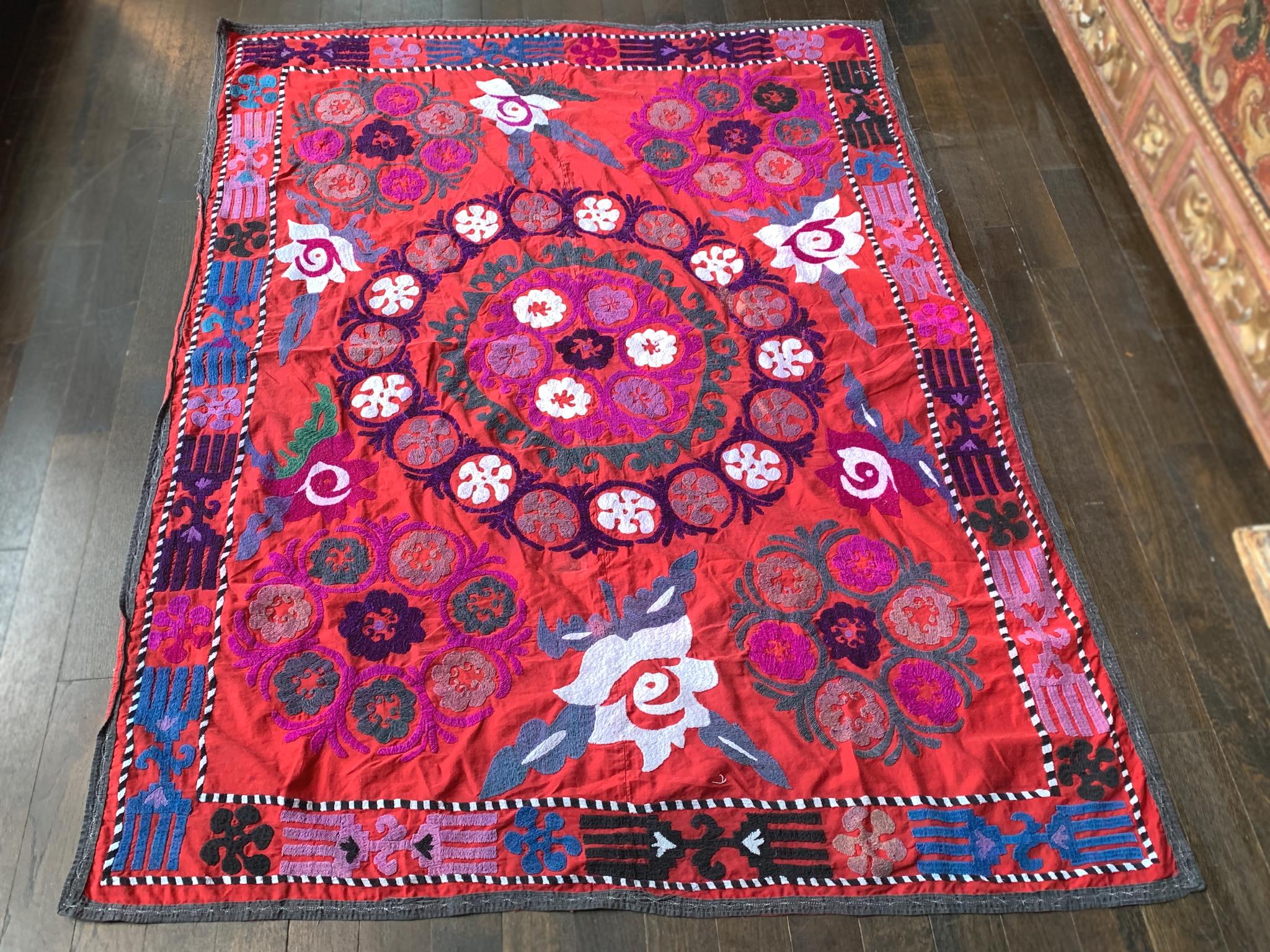 A lovely Suzani hand sewn and hand-embroidered in the 20th century. It's comprised of a deep red base fabric with embroidery in a palette of pink, blue, green, purple, black, and white. A traditional floral design populates the central field which