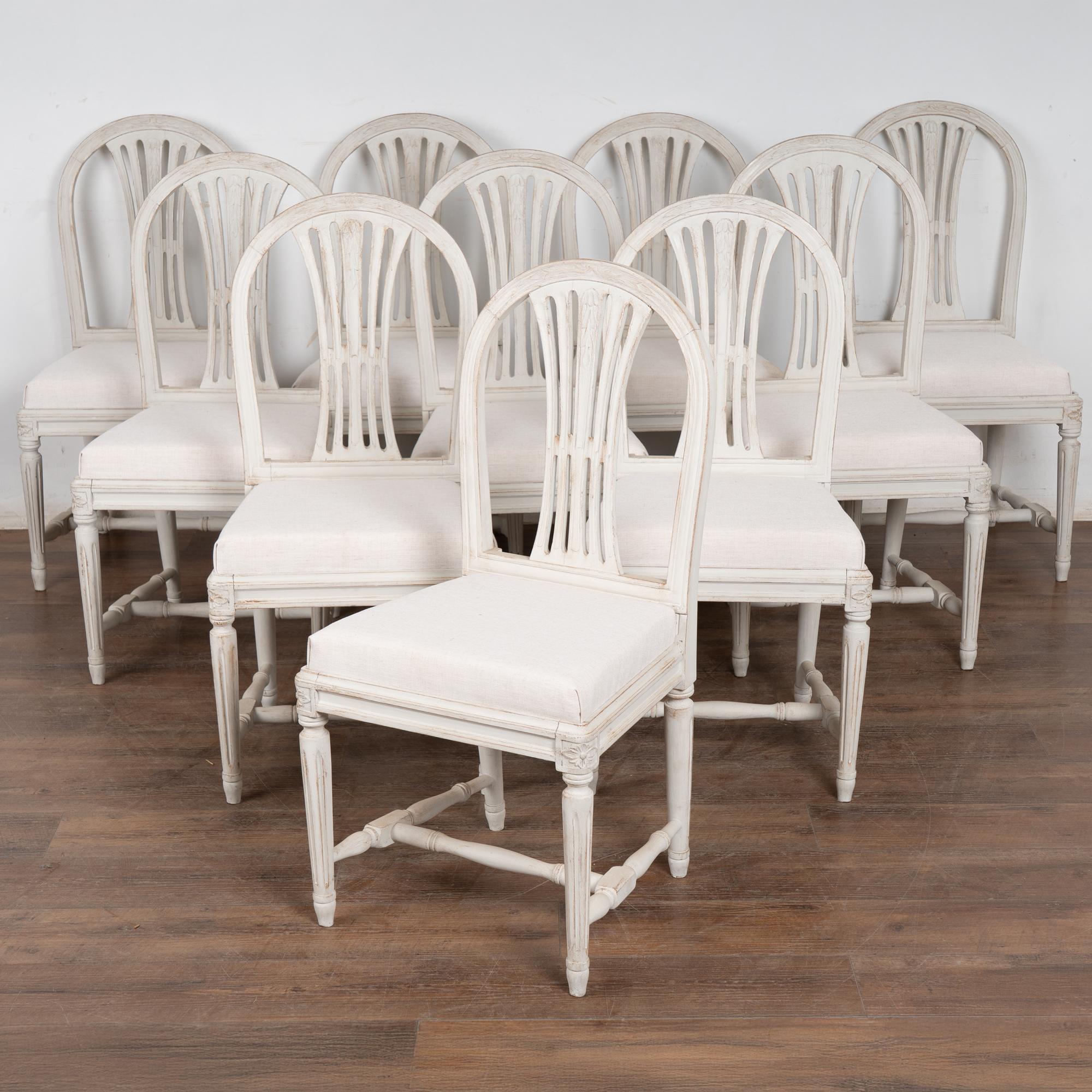 Set of 10 dining chairs from the early 1900's. Newer white painted finish accents the Swedish styling with carved details along the arched back, and floral motif topping the turned and fluted legs. 
Restored, the chair frames are strong and stable,