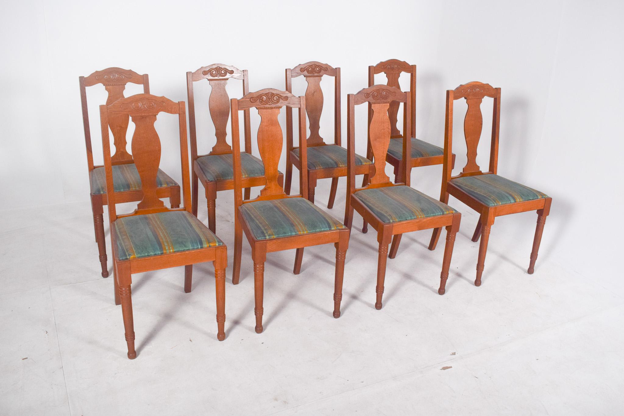 Conjunto de ocho sillas de roble. Asientos tapizados en tela de rayas en tonos verdes, azules y amarillos, con una estructura firme y bien conservada. El respaldo presenta un diseño recortado con un motivo central en forma de espiral estilizada,