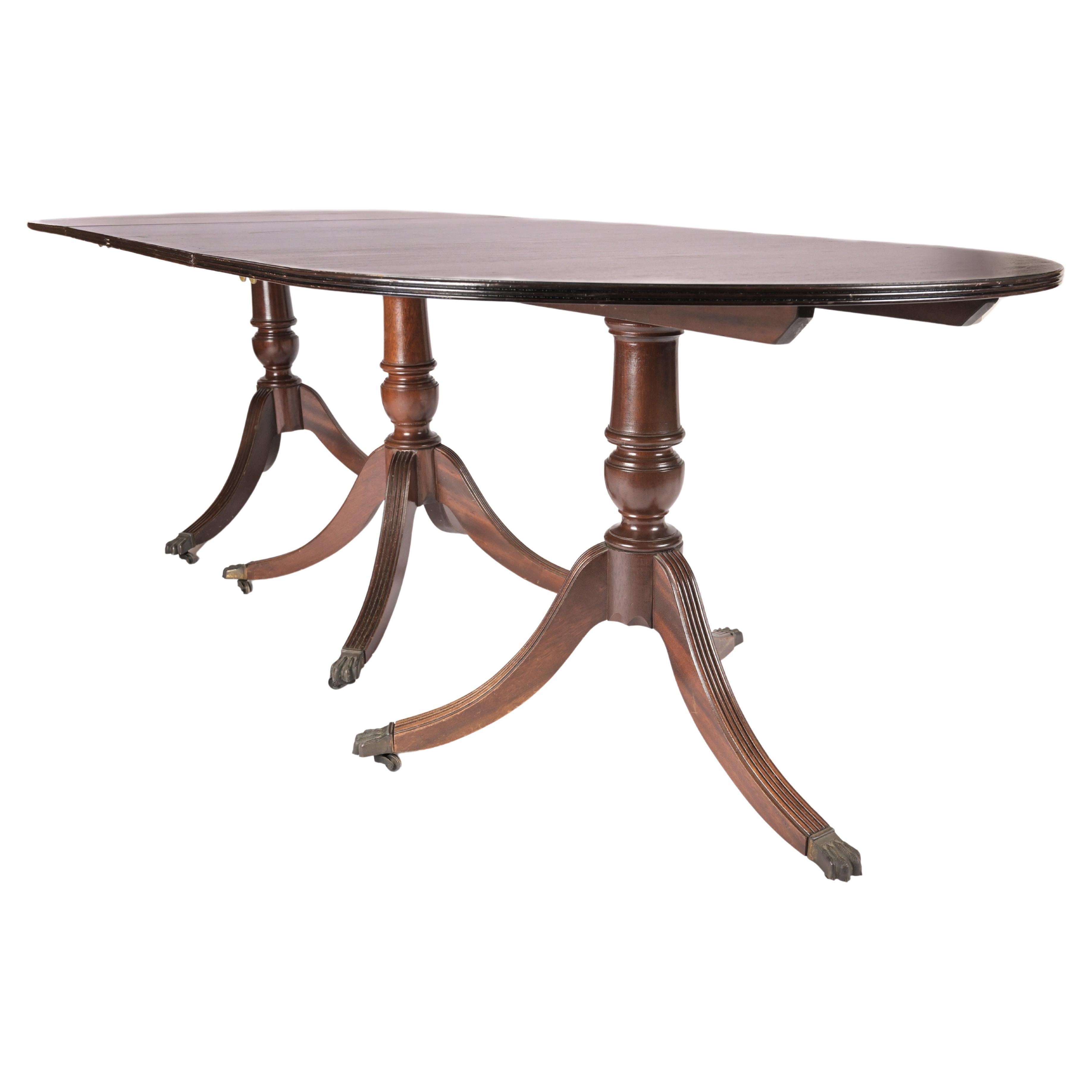 An early 20th century English Sheraton Style Mahogany pedestal base dining table with 3 pedestals having splayed legs with brass lions paw feet and casters. The top of each of the three sections hinges on the pedestal base for storage with two