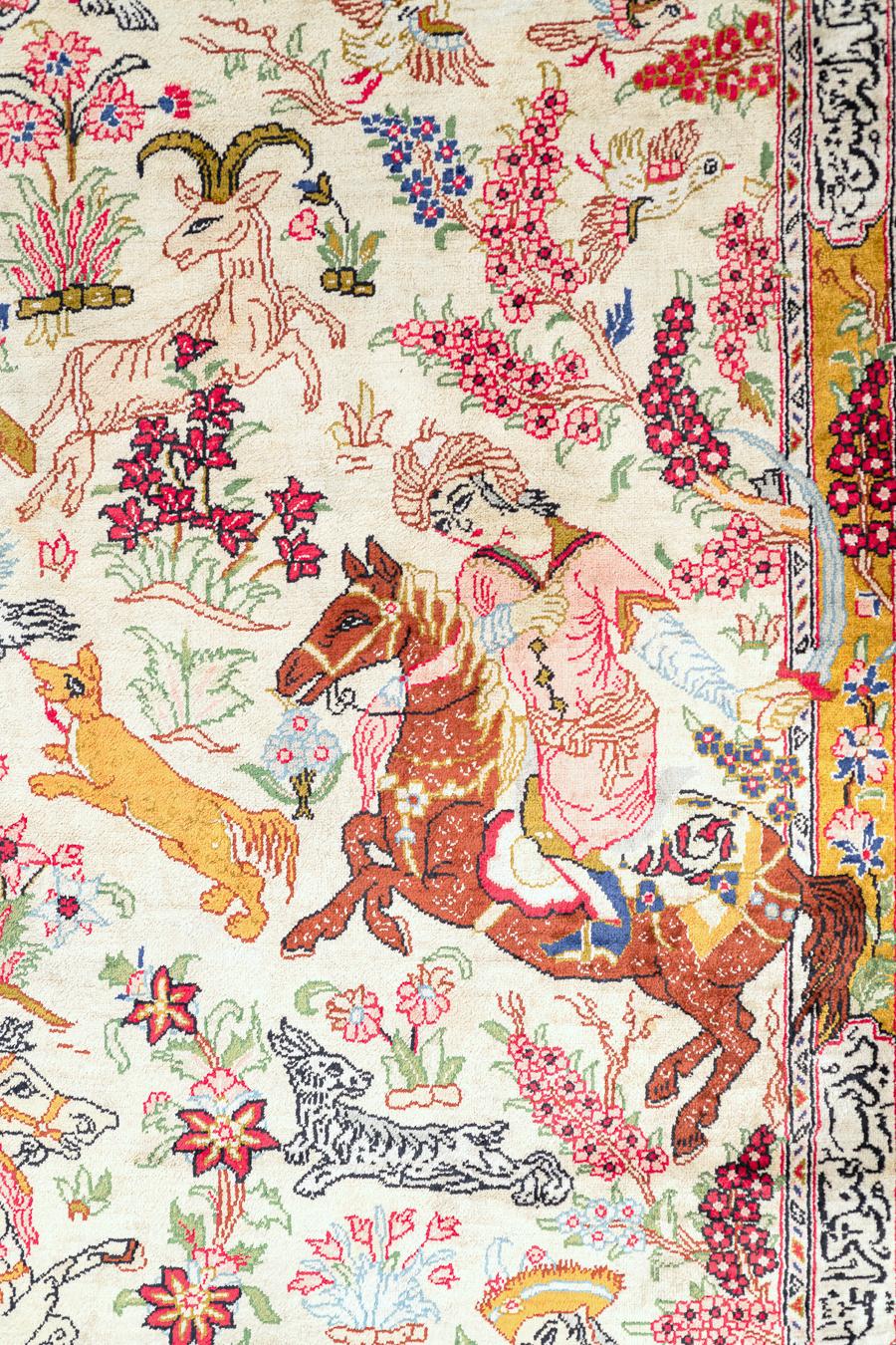 Ce tapis en soie du XXe siècle de style Ghom présente un récit captivant tissé dans son dessin : une scène de chasse dynamique avec des cavaliers, des animaux et une végétation florissante disposés sur un champ de soie chatoyant. Chaque élément, du