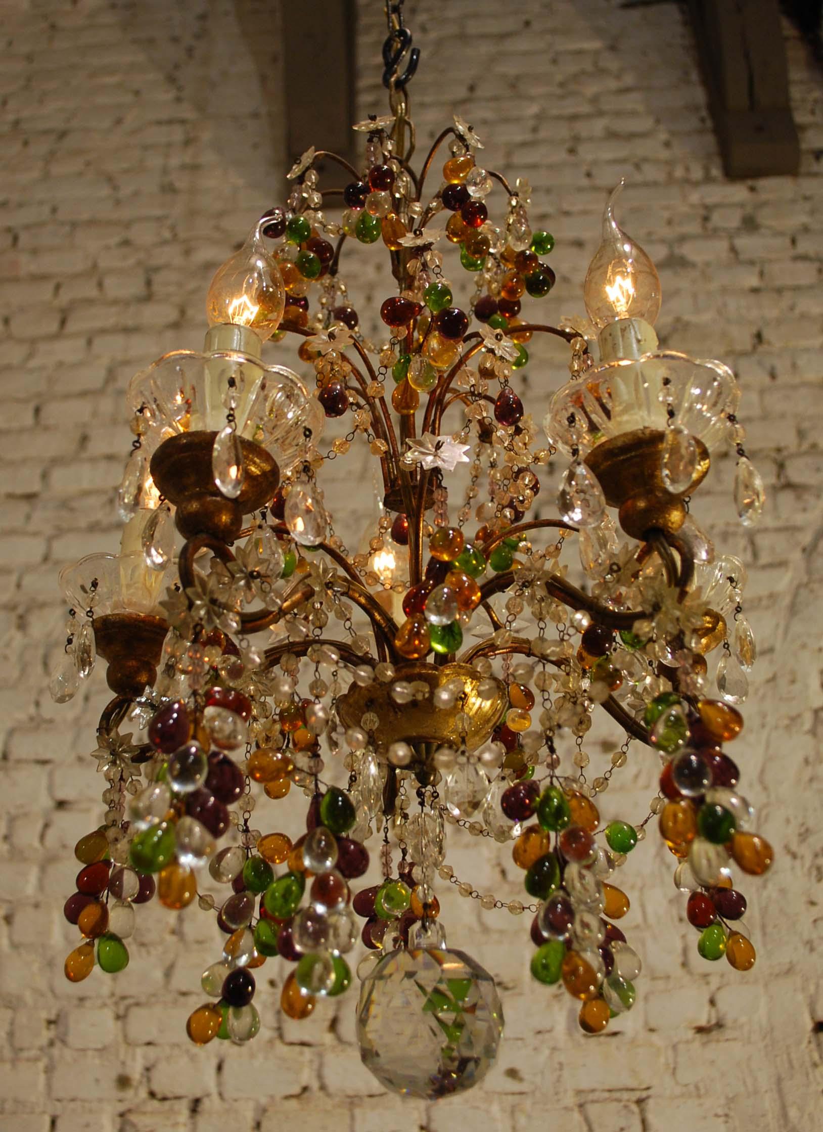 This small but elegant Italian chandelier has five lights. It has a forged steel frame that is gold gilt.
The arms have crystal bobeches on top of giltwood posts. It has adorning crystal multicolored grapes with macaroni bead swags and stars