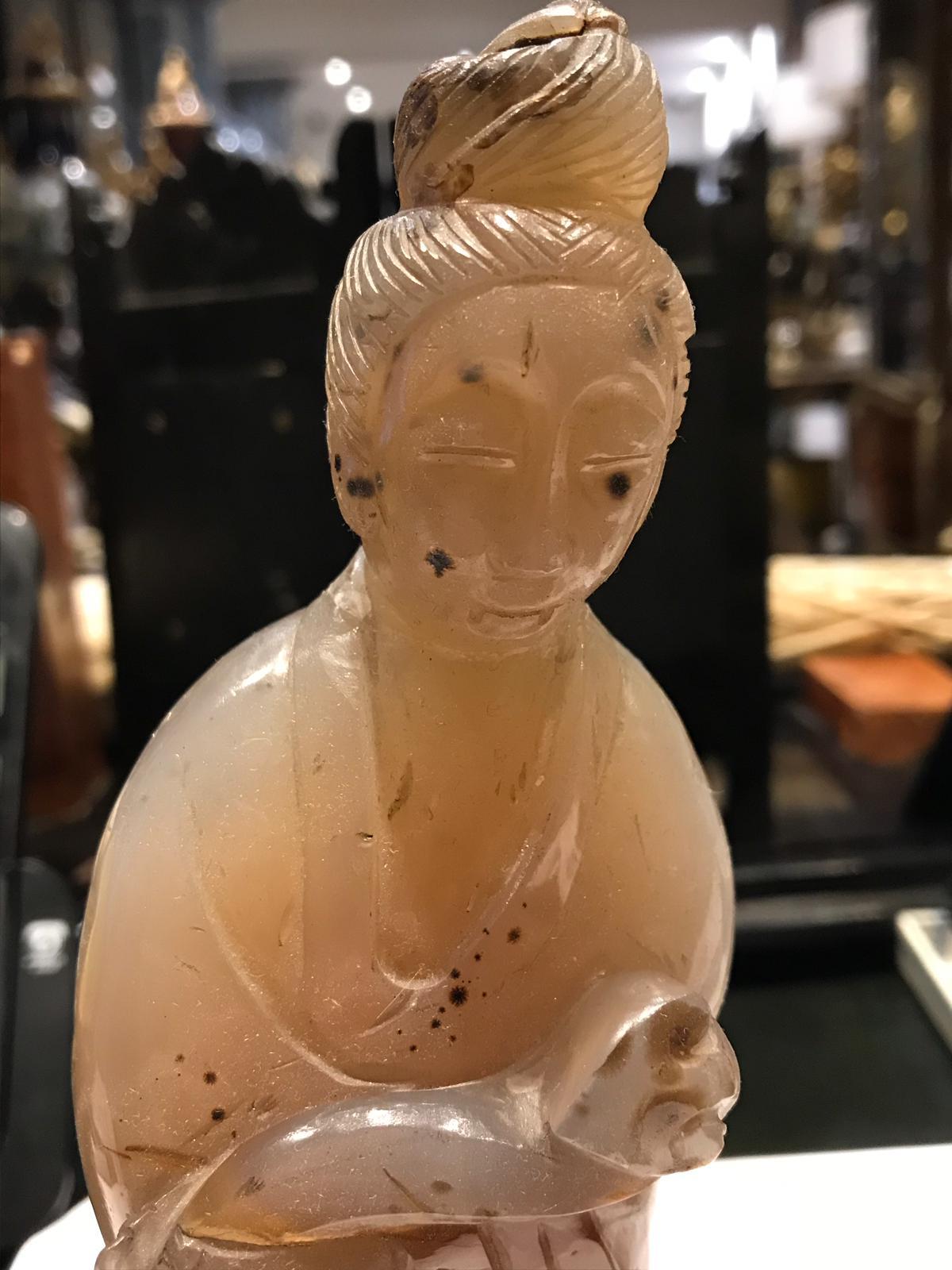 A smoky agate carved figure of an Asian Woman. One of her hands is cusping something that lies across her figure. She has long robes in traditional dress and wears a bun. The stone is mostly smooth with some dents and scratches, but nothing drastic.