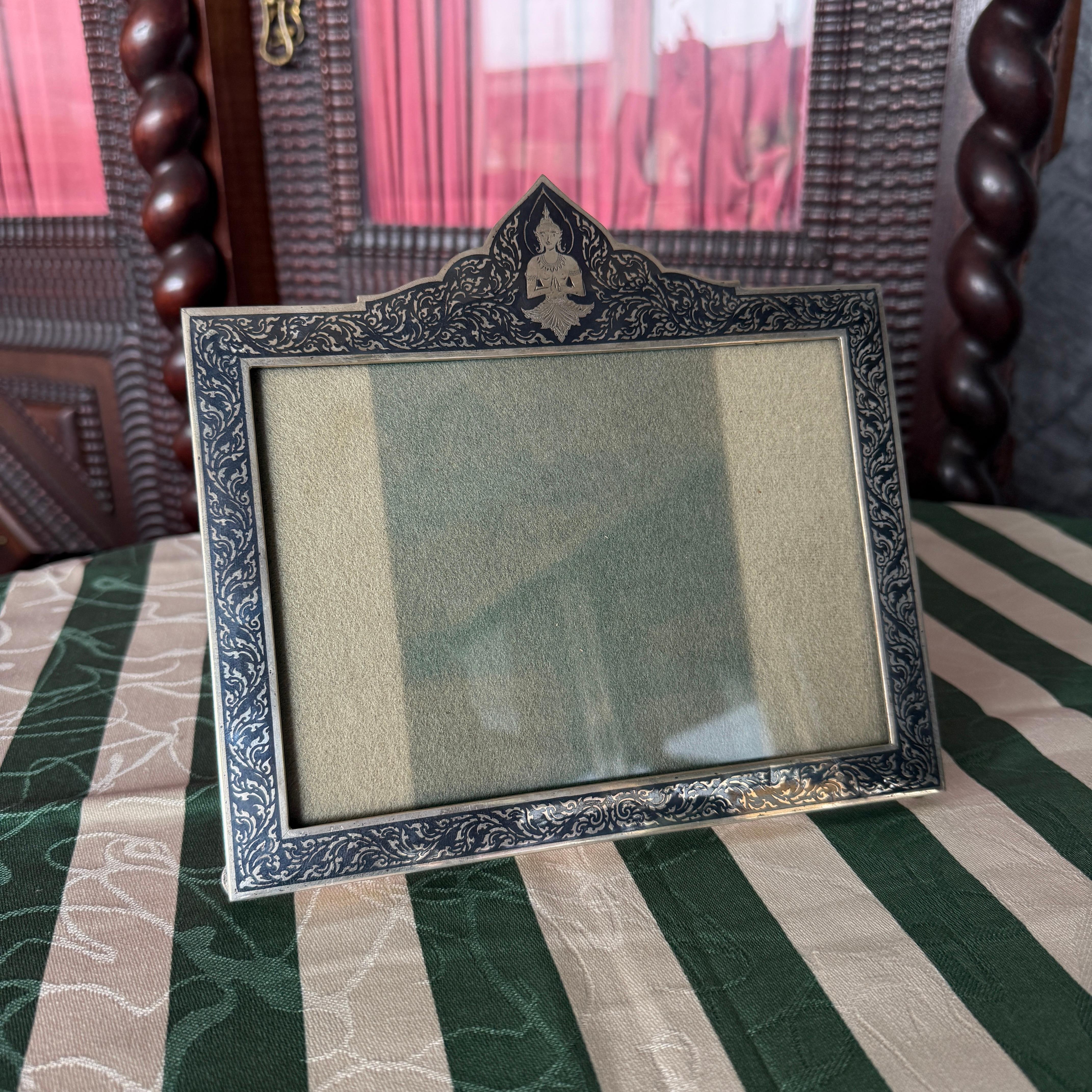 This elegant 20th Century Thai picture frame showcases the refined artistry of traditional silverwork combined with the striking contrast of niello inlay. Handcrafted in sterling silver, the frame features a rectangular form adorned with intricate