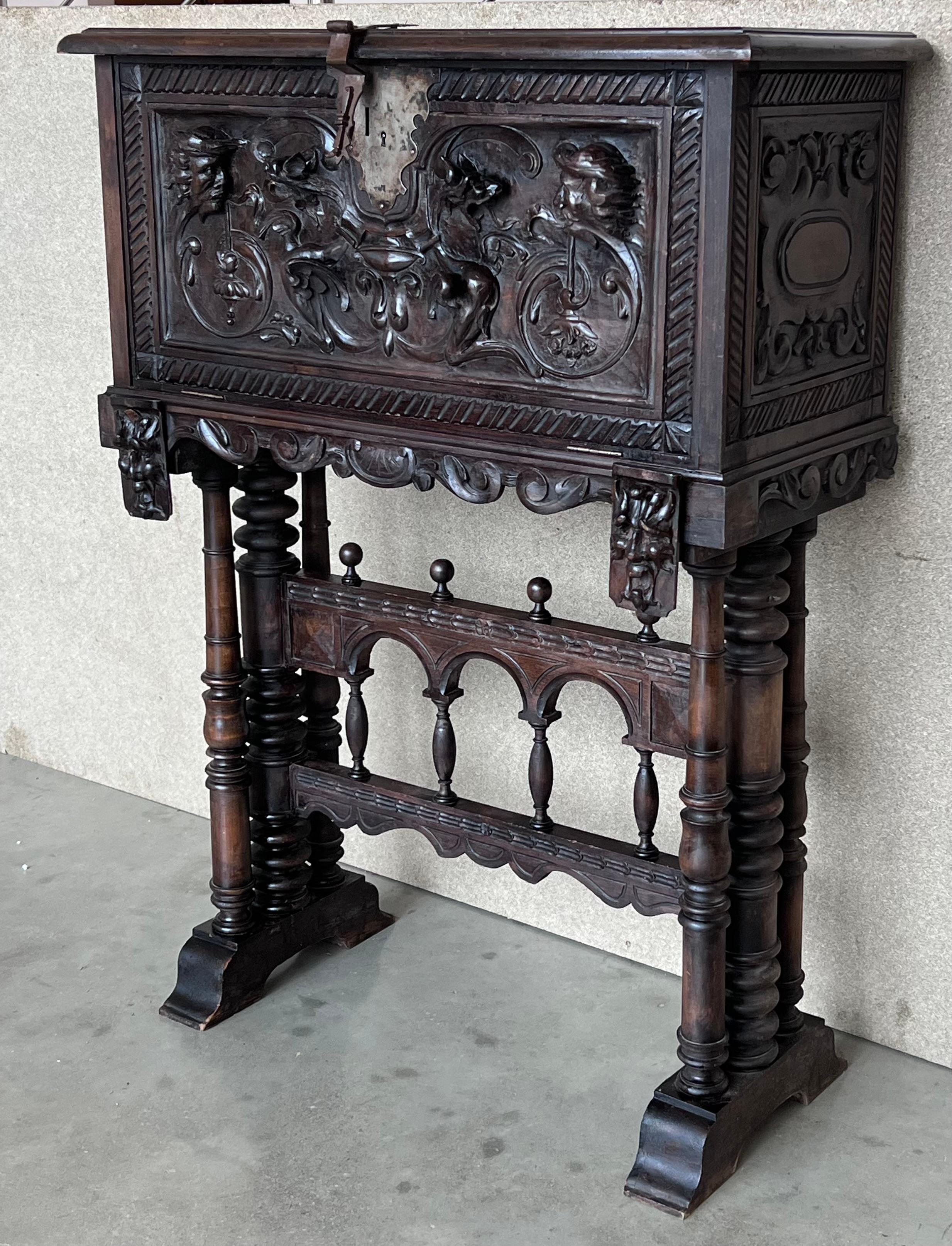 This Bargueno is embellished with a carving of Don Quixote de la Mancha bordered with branches of leaves. The desk is supported by a pedestal joined by a bridge of turned columns. The front legs contain a carving of two wind god heads with an