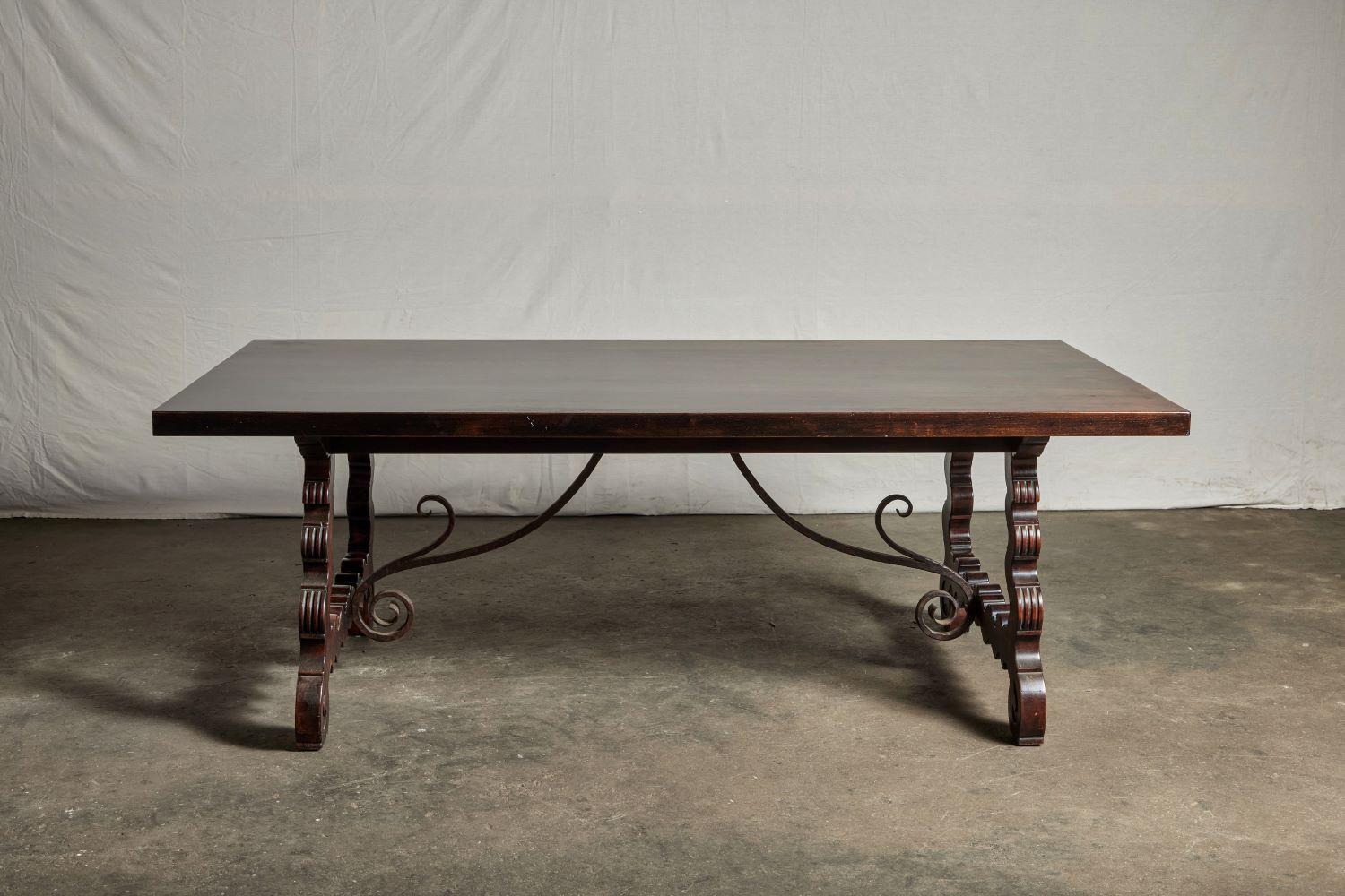 The dining table is made of mahogany and brass and features a slab top on a trestle base with iron brackets and scroll cut legs held together by an S-curve brass detail.