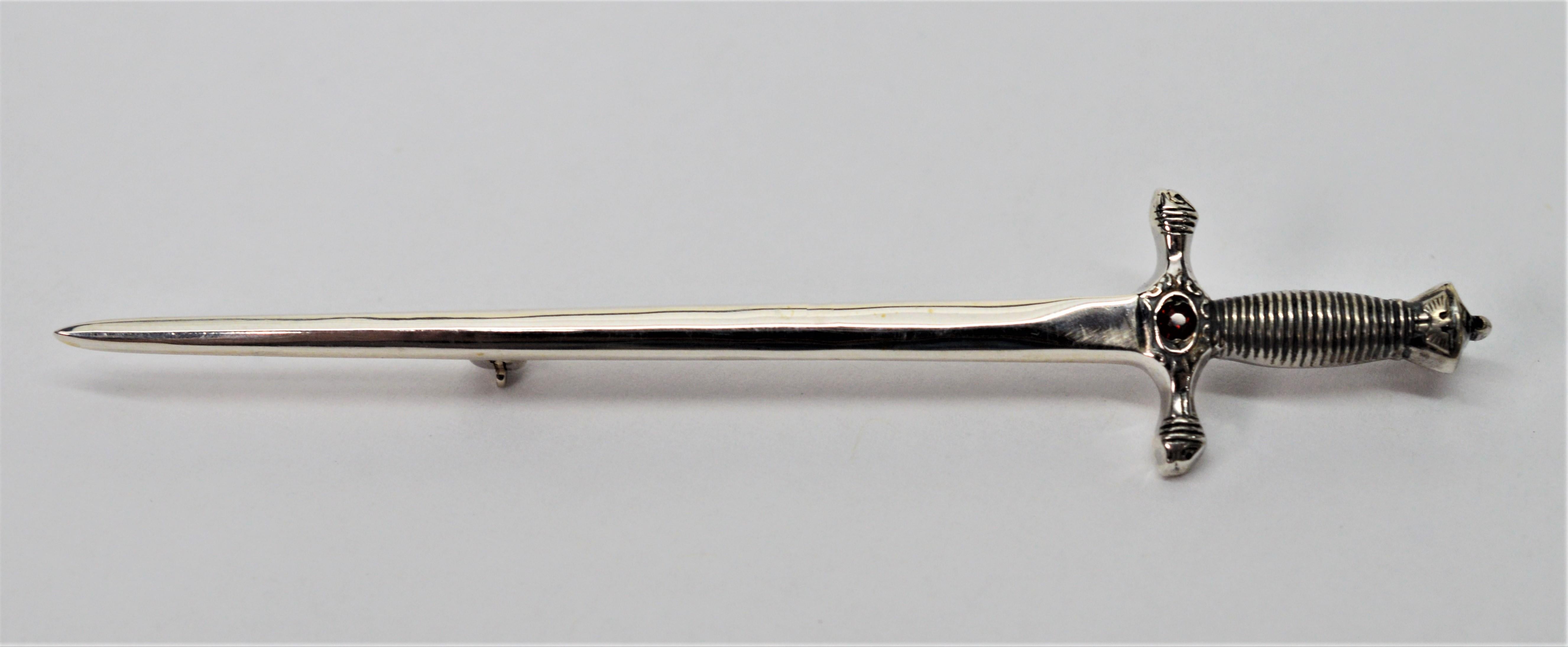 Wield power when wearing this sword pin. Made in sterling silver with a garnet gemstone on the guard. Crafted in detail with a textured grip and decorative pommel. Measures 2-7/8 inches in length. The pin closure is a modern safety catch. In gift
