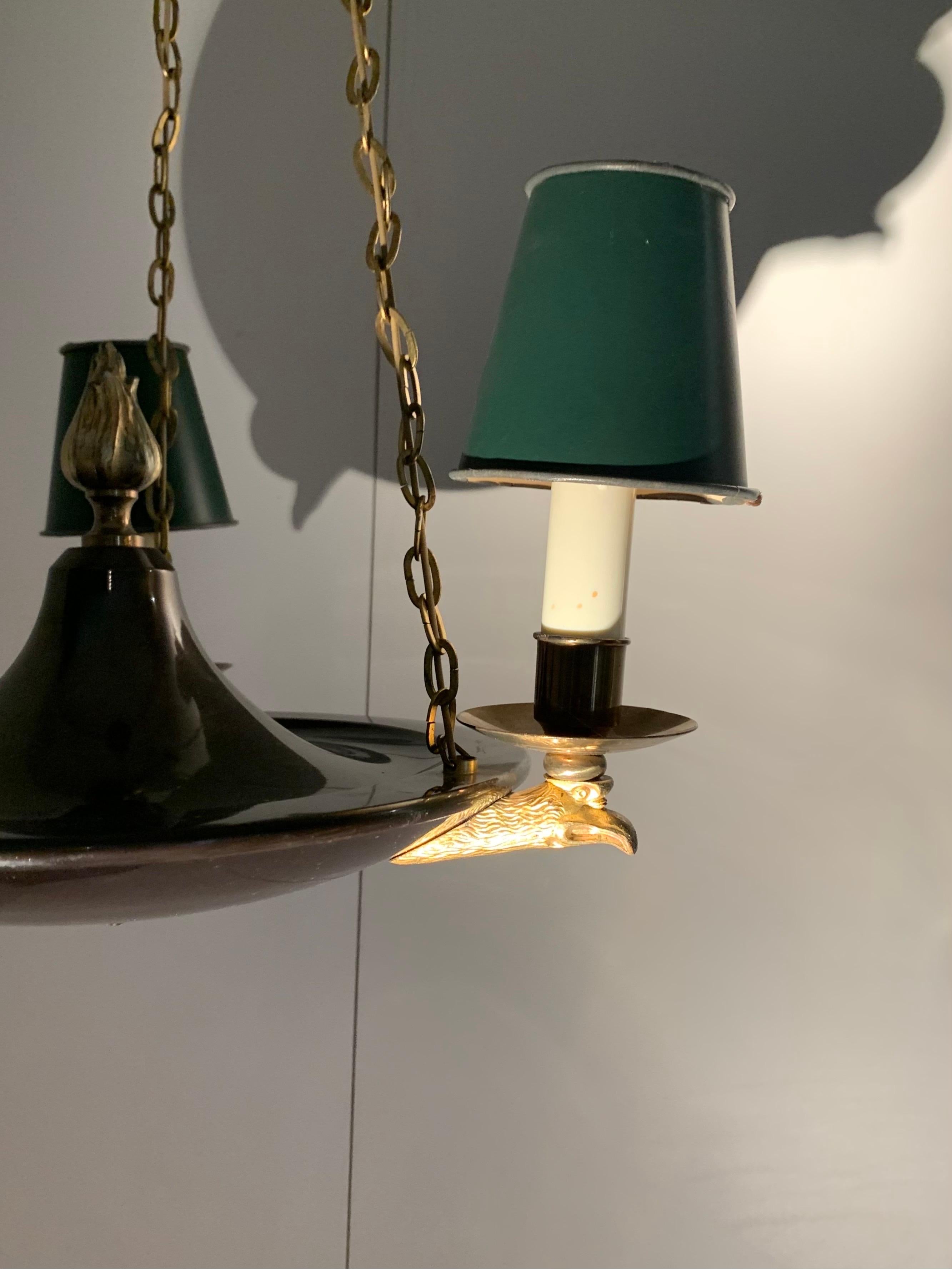 Una lampada da soffitto in stile impero, in metallo laccato e ottone, con tre punti luce che poggiano su teste d'aquila e rifiniti con una fiamma; i tre punti luce sono rifiniti con piccoli paralumi originali verdi.